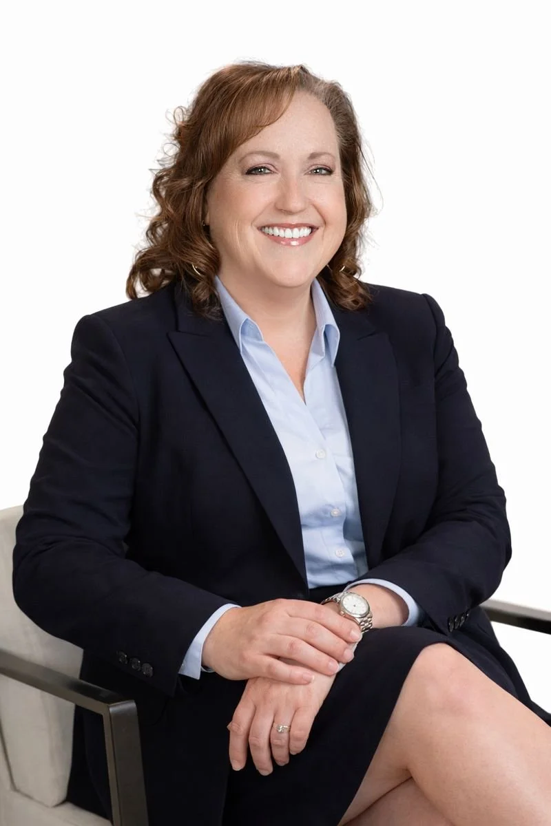 A woman with red hair wearing a navy blazer, light blue shirt, and a silver watch, sitting on a white chair and smiling.