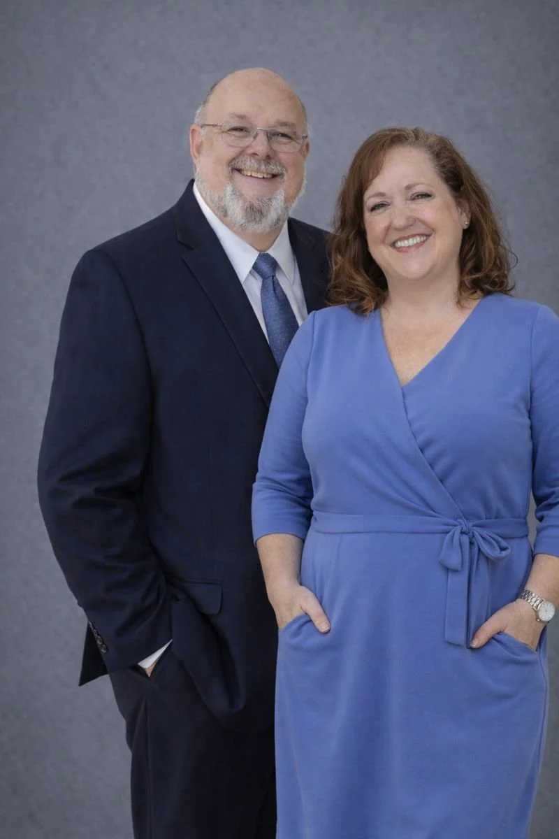 A smiling middle-aged man in a dark suit with a light blue tie standing next to a smiling middle-aged woman in a blue dress with her hands in her pockets, against a plain gray background.