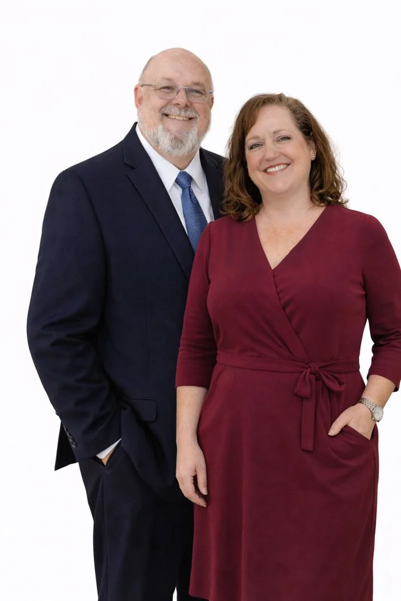 A smiling man with glasses, a beard, and a suit, standing next to a smiling woman with shoulder-length hair and a red dress, against a white background.