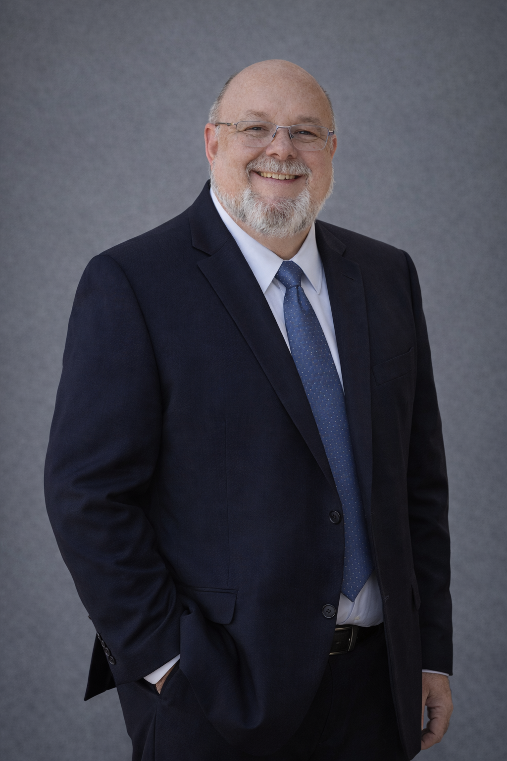 A smiling middle-aged man with glasses, a beard, and a bald head, wearing a dark suit, light blue shirt, and blue tie, standing against a plain gray background.