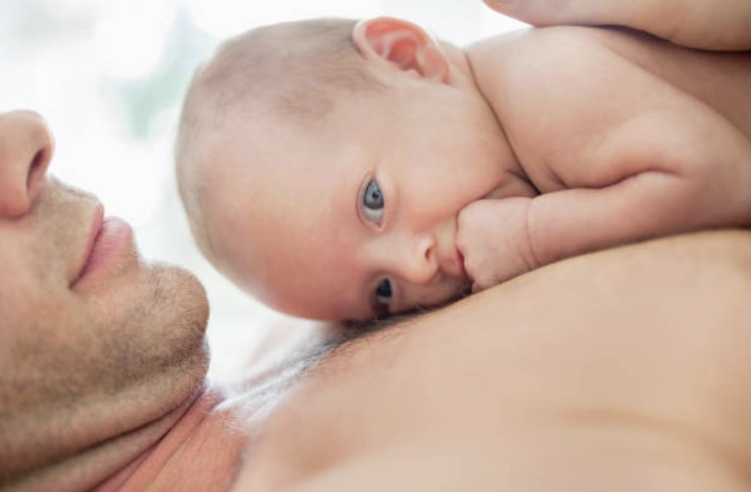 A newborn baby nursing on a man's chest, with the man lying down, in a bright, soft-lit setting.