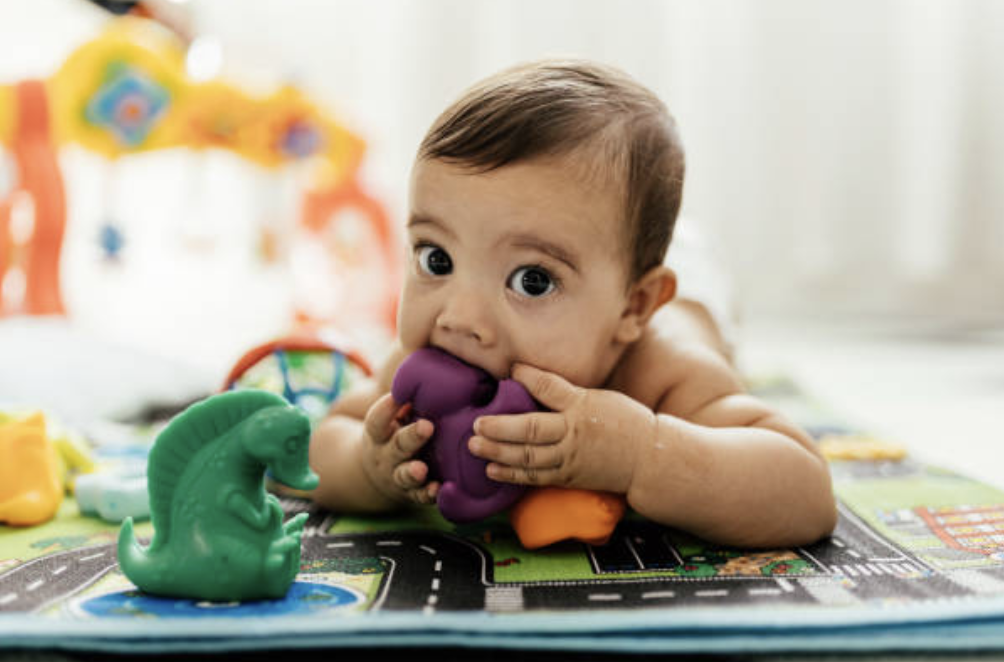 A baby lying on a play mat, holding a purple toy and licking it, surrounded by colorful toys.
