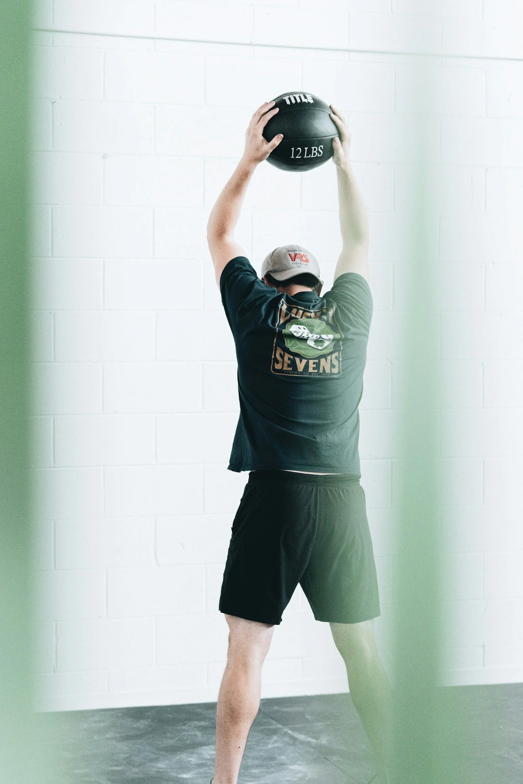 A man exercising indoors, holding a 12-pound medicine ball overhead. He is wearing a gray cap and black athletic shorts, with a black T-shirt that has a logo on the back. The background features a white brick wall.