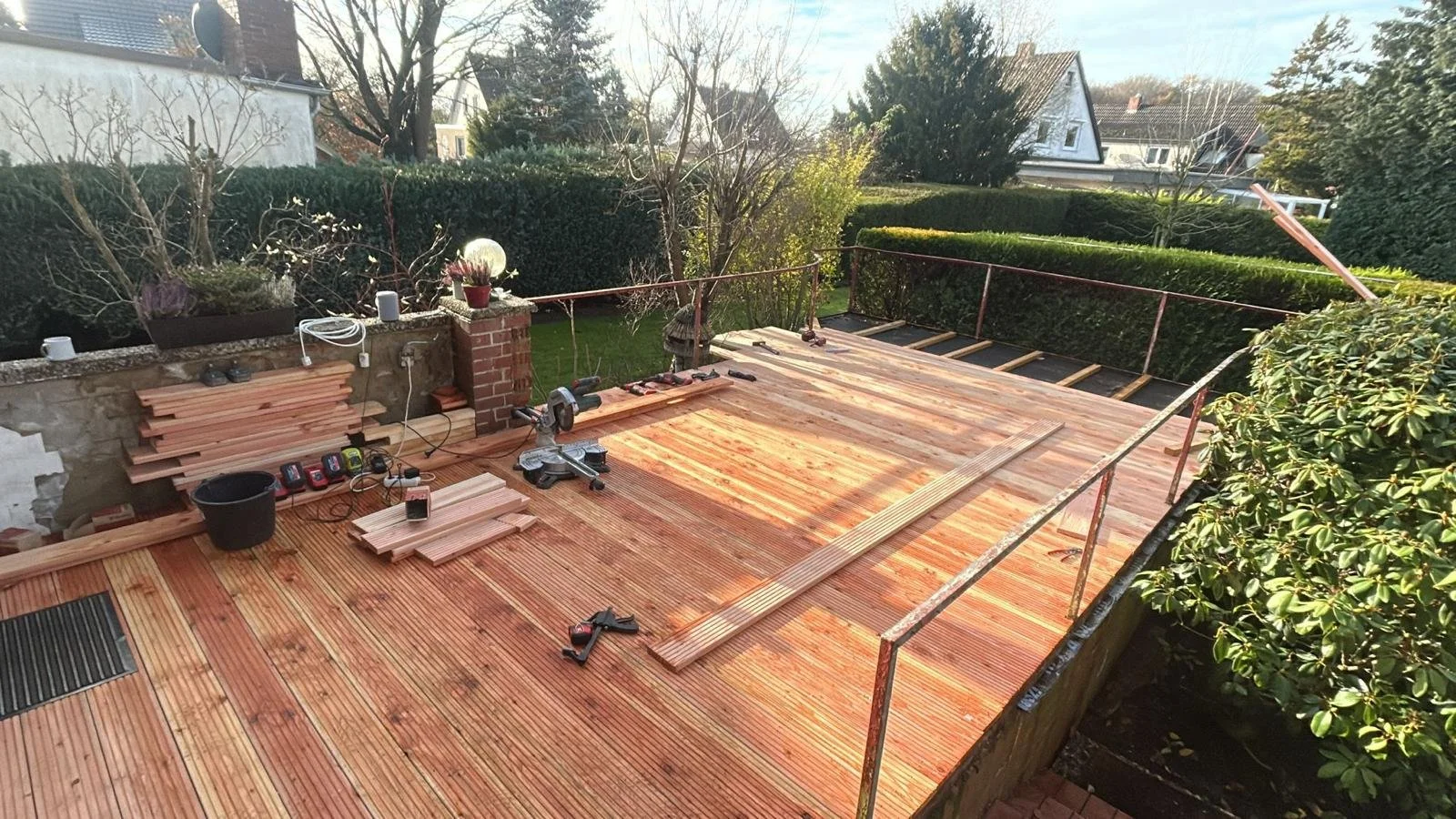 Garten im Bau, auf dem Boden werden Holzdielen verlegt, verschiedene Werkzeuge und Holzstücke liegen bereit, im Hintergrund Bäume und Hecken.