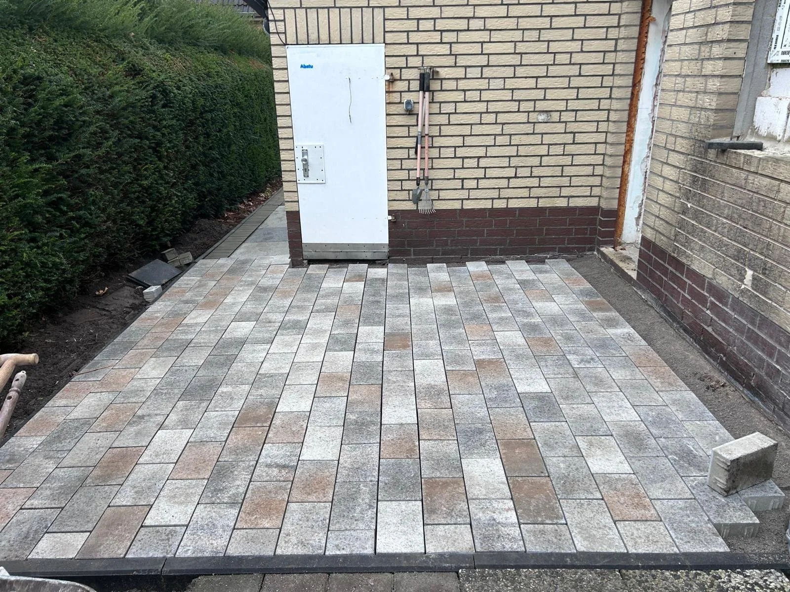 Neue gepflasterte Terrasse aus bunten Ziegelsteinen vor einem Haus mit einer Hecke auf der linken Seite.