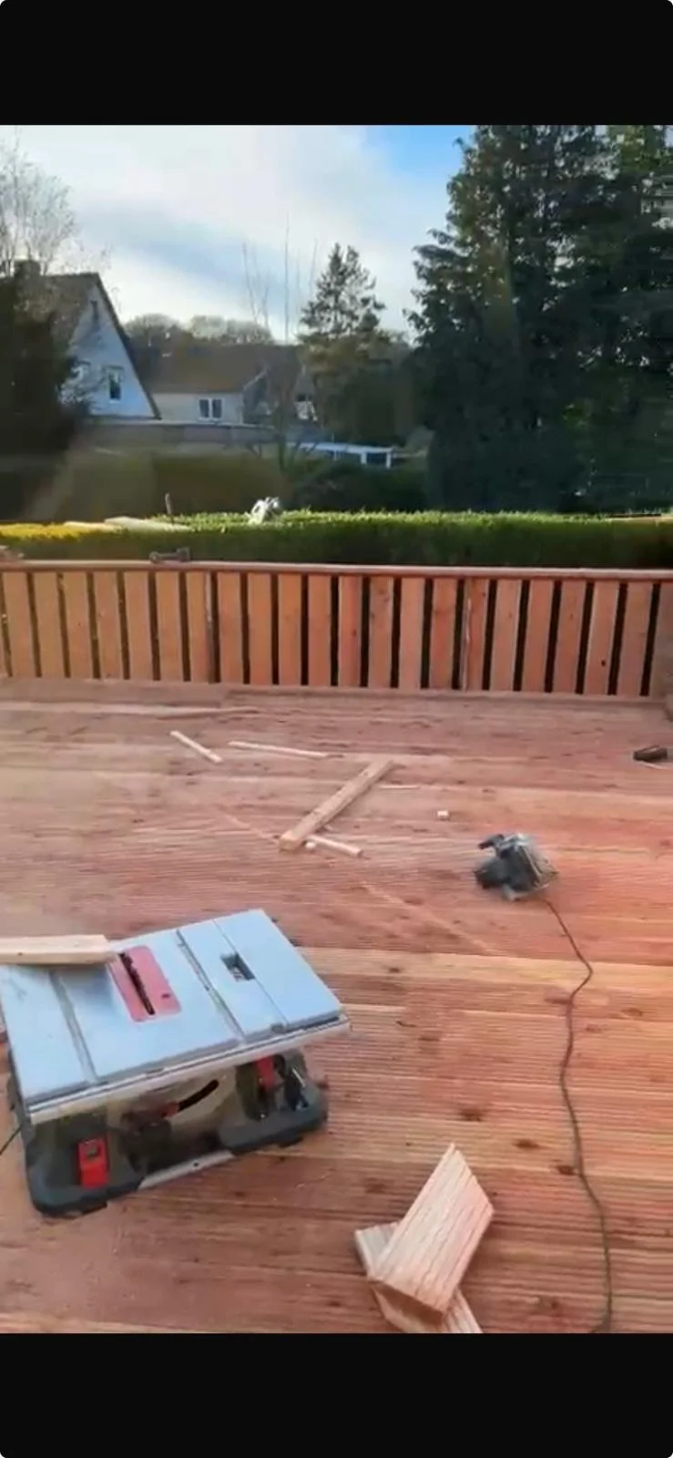 Holzterrasse im Bau mit Werkzeugen und Baumaterialien, Blick auf einen Garten mit Büschen und Bäumen unter einem bewölkten Himmel.