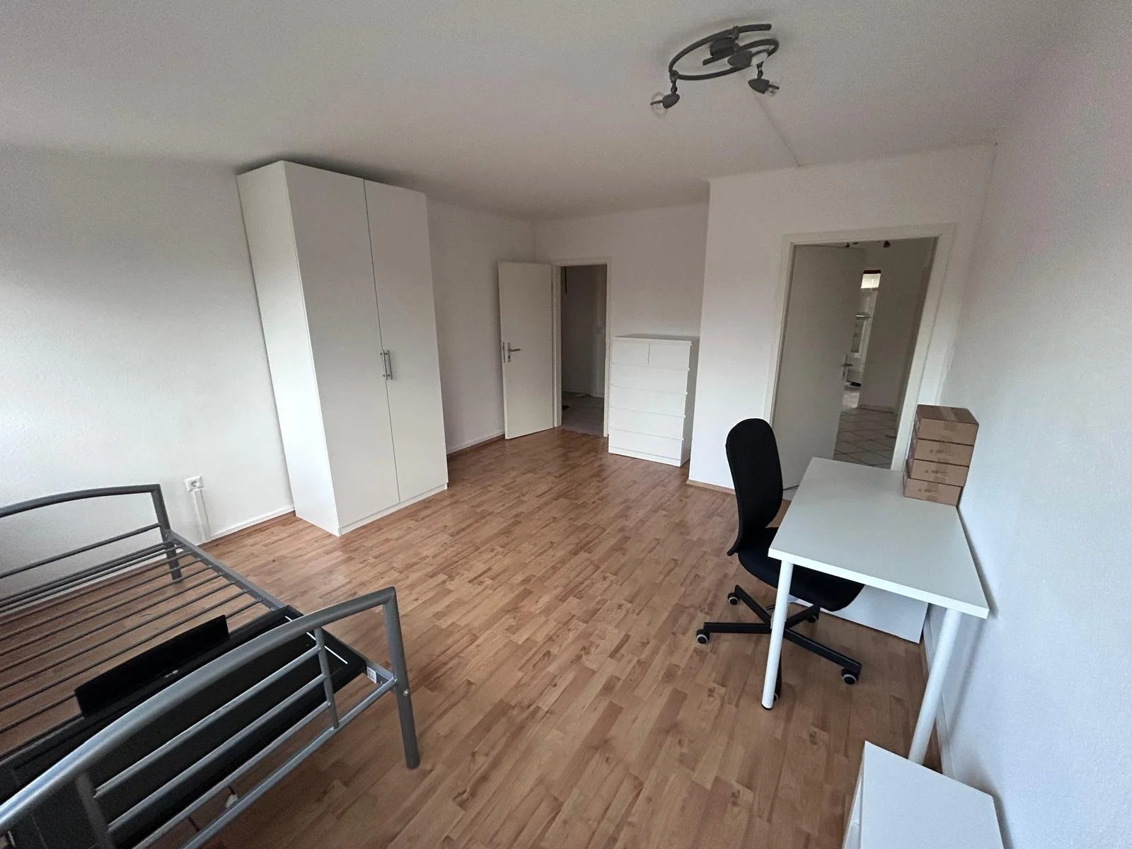 Leerraum mit Holzboden, weißem Schrank, Bettgestell und Schreibtisch mit Stuhl, an der Decke eine Lampe.