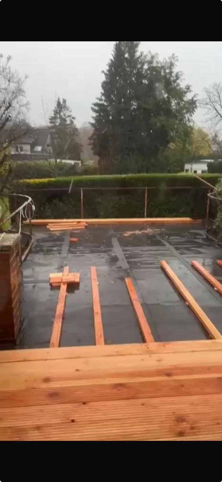 Balkon mit Holz-Dielen, die verlegt werden, im Regen, im Hintergrund Bäume und Häuser.
