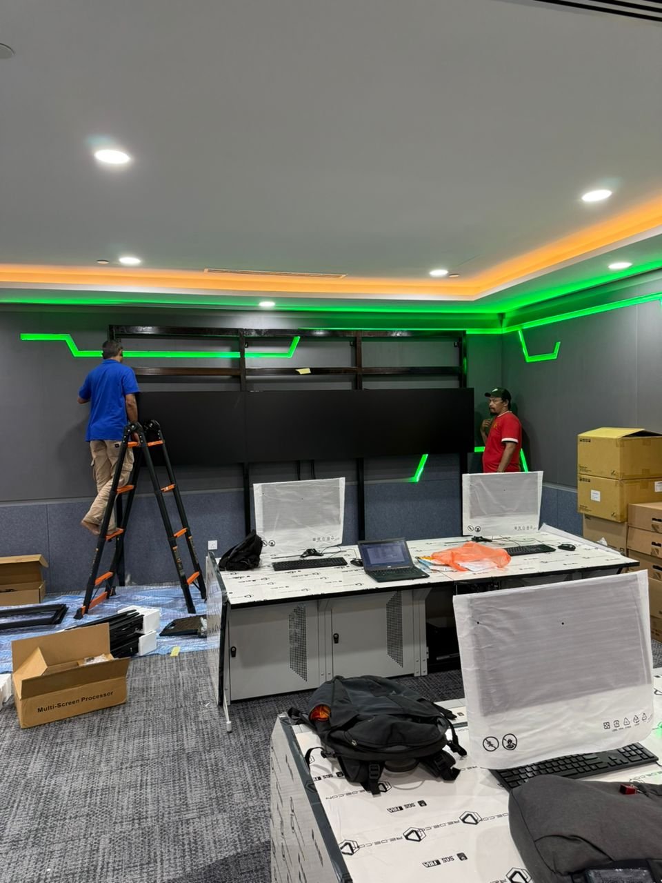 Two workers assemble a wall with green LED strip lighting in a modern office or control room. One worker stands on a ladder, and the other stands on the floor. Desks with computers and monitors are in the foreground, with packaging and boxes around.