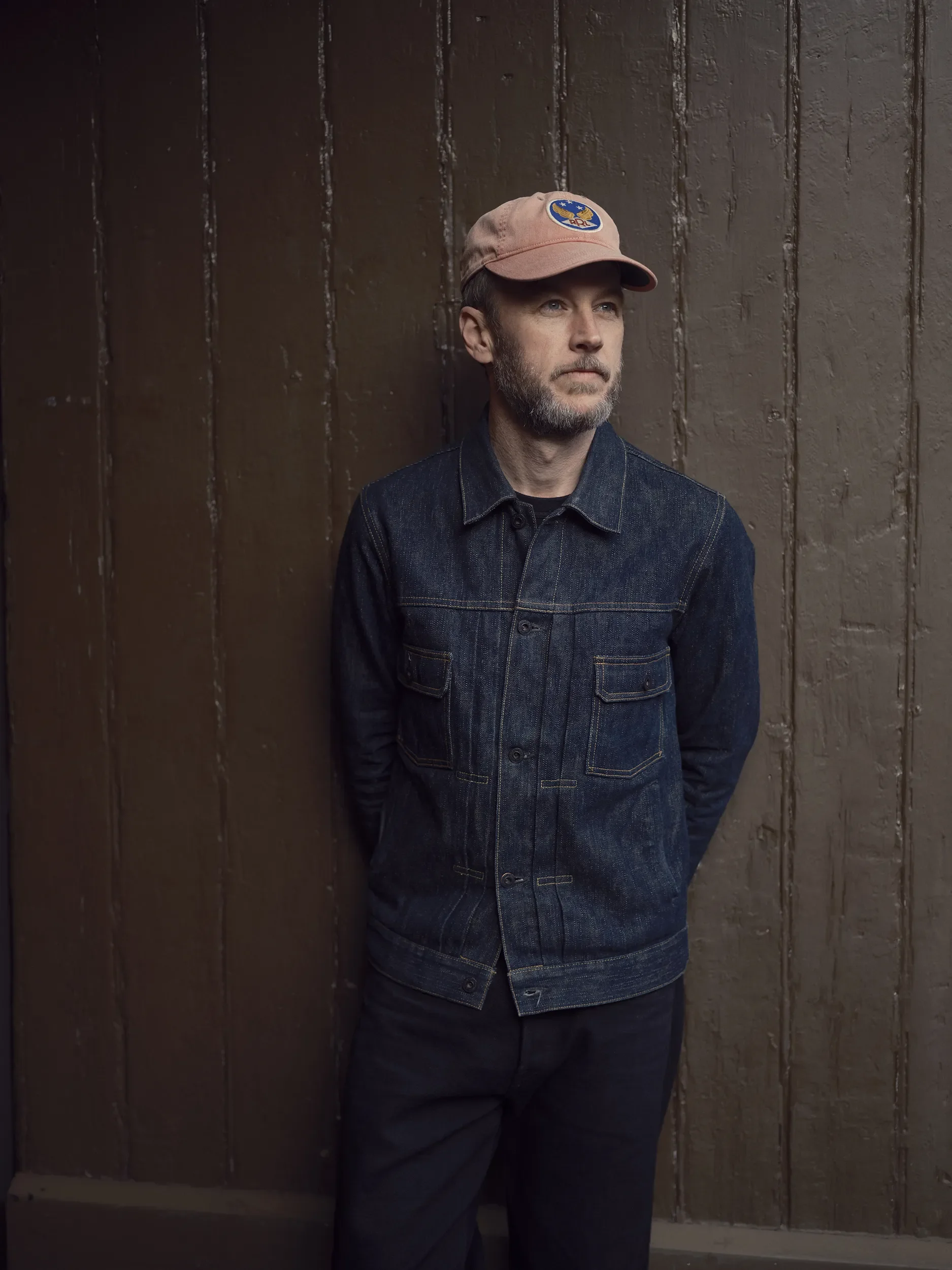 A man with a beard wearing a light pink cap with a NASA patch, a dark denim jacket, and black pants standing against a brown wooden wall.