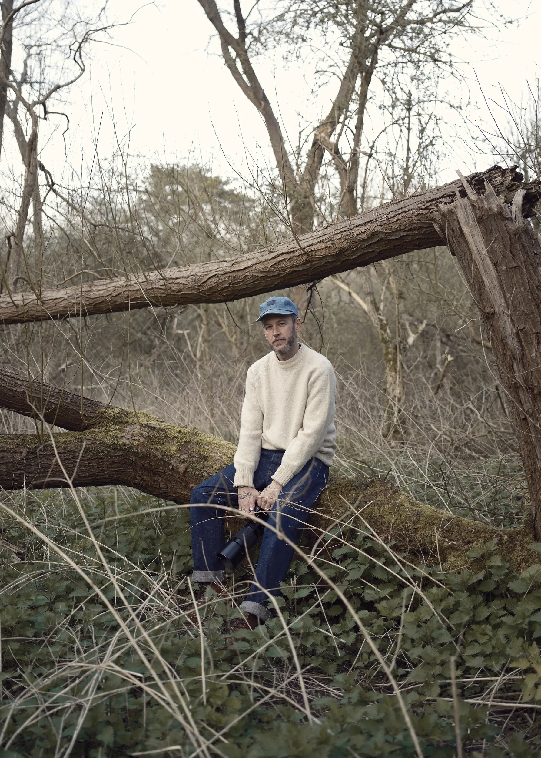 A man wearing a beige sweater, blue jeans, and a blue cap sits on a fallen tree in a dense, leafless forest. He is holding a large camera lens and looking at the camera.