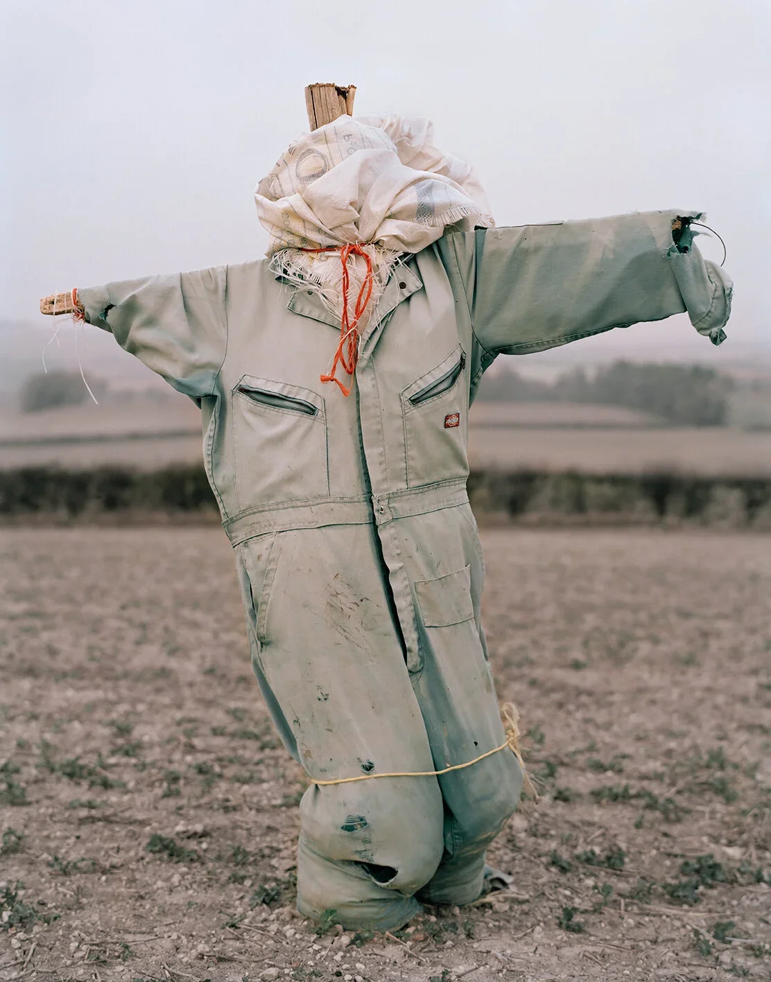 A scarecrow dressed in an old, weathered work jumpsuit with a head made of a wooden block wrapped in cloth, standing in a barren field.