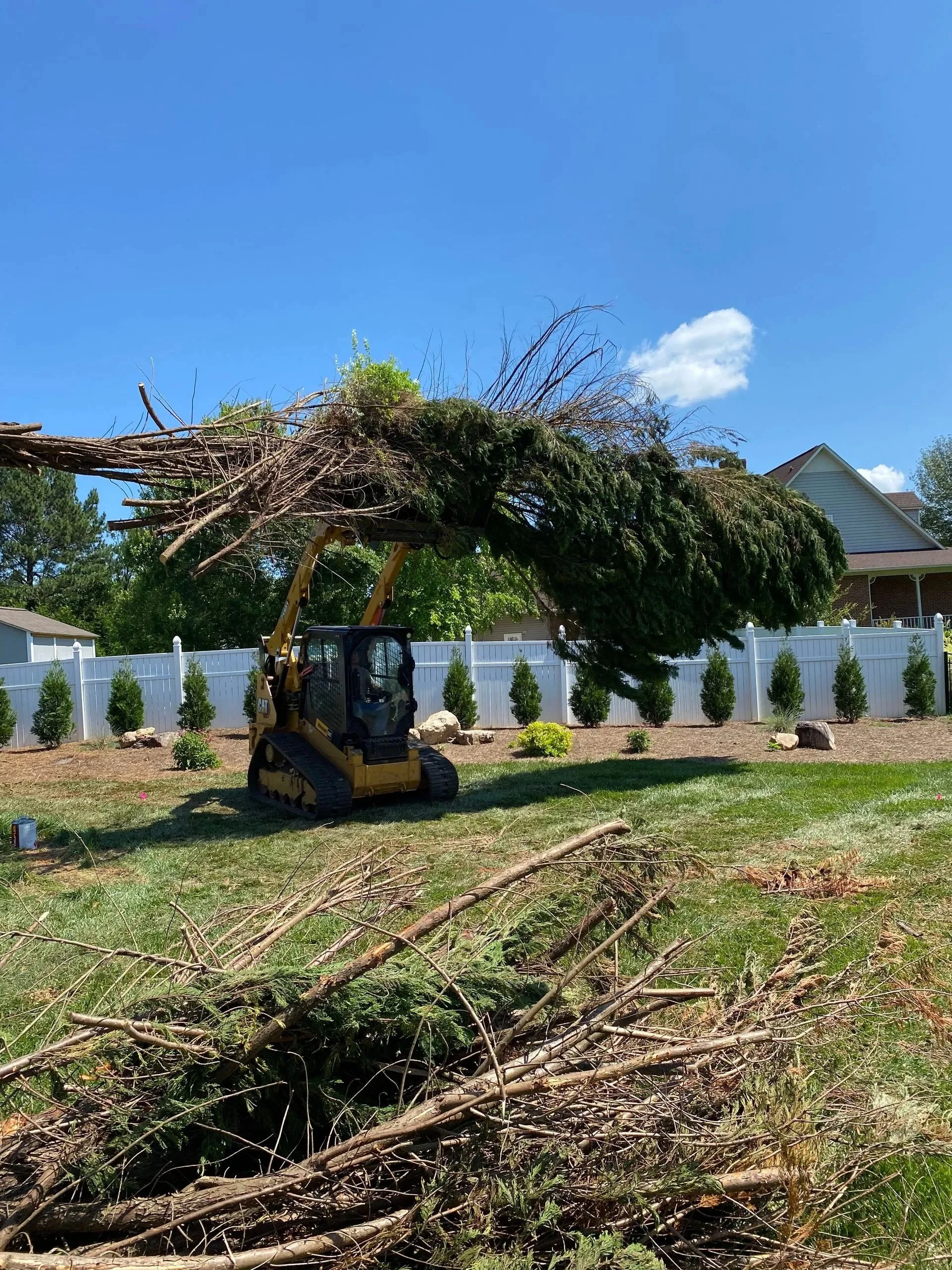 A small bulldozer lifting a fallen tree branch in a backyard with a white fence and small trees.