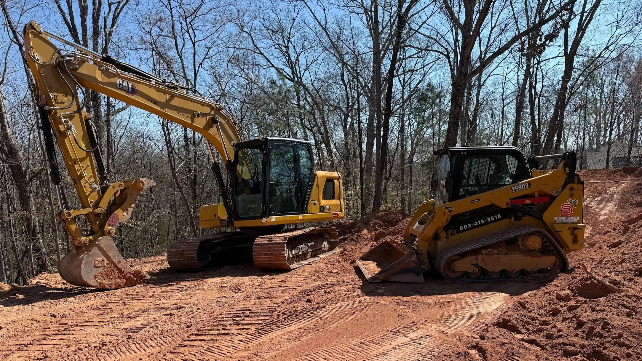 Two yellow excavators working on a dirt construction site surrounded by leafless trees.