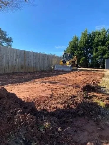A construction site with a backhoe loader working on a cleared dirt area, fenced yard, trees, and a blue sky with clouds in the background.