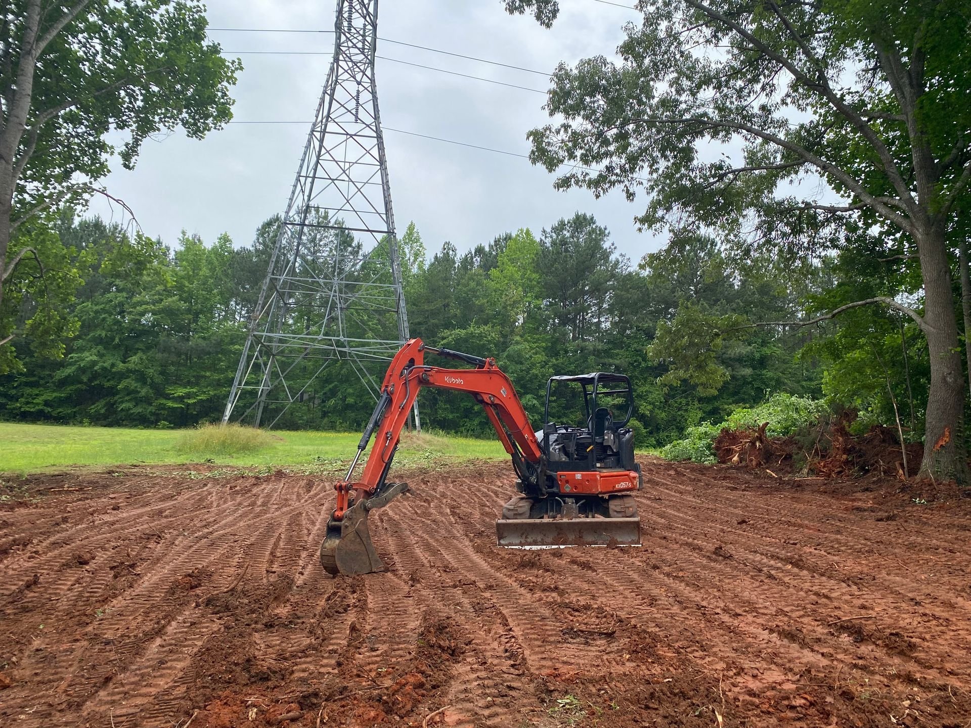 Construction site with a small orange excavator on reddish dirt, surrounded by green trees and a metal transmission tower in the background.