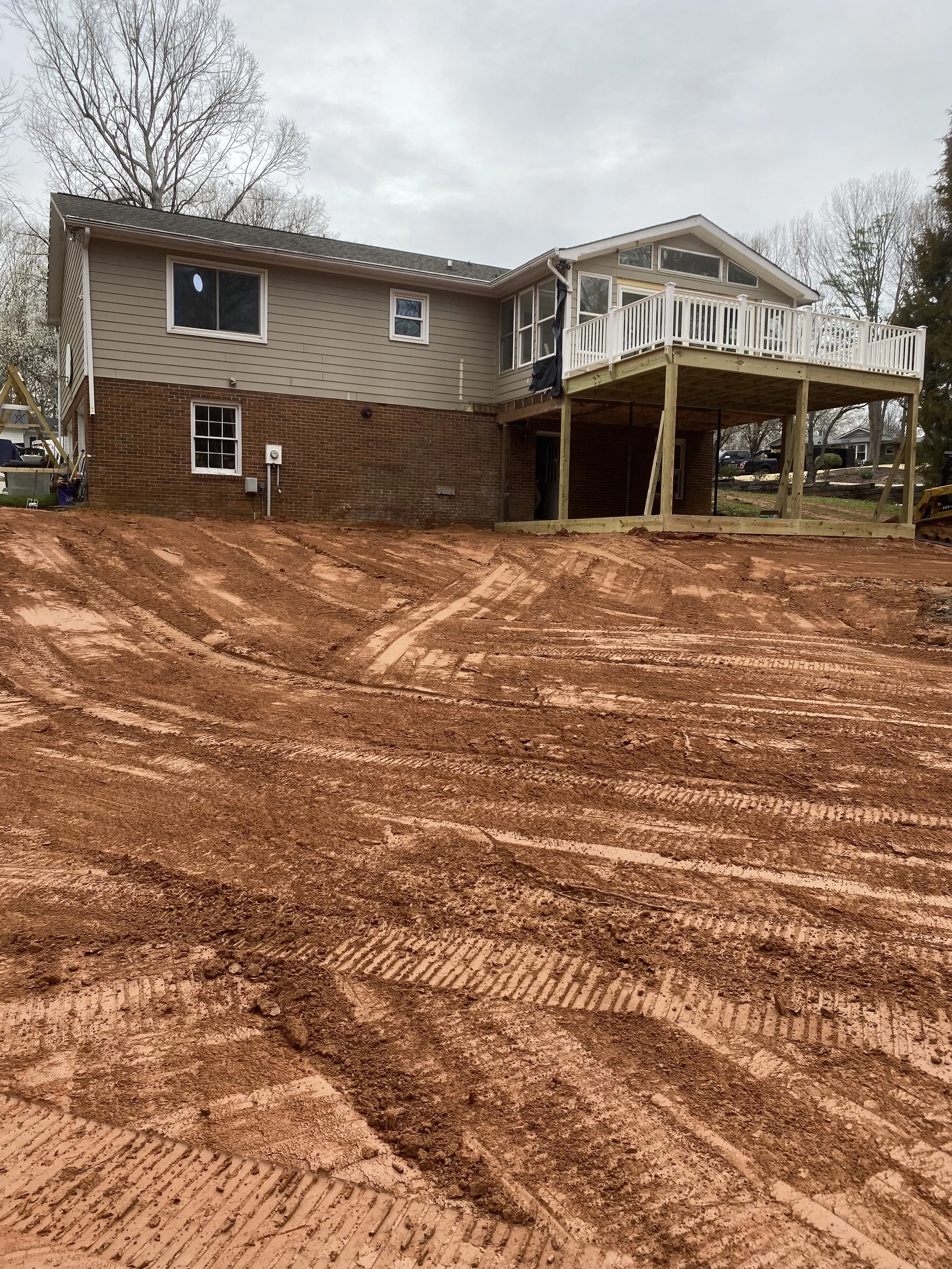 House with a brick lower level and beige siding upper level, featuring a wooden deck with railing expanded from the house. The backyard is under construction, with dirt and tire tracks from construction equipment visible in the foreground.