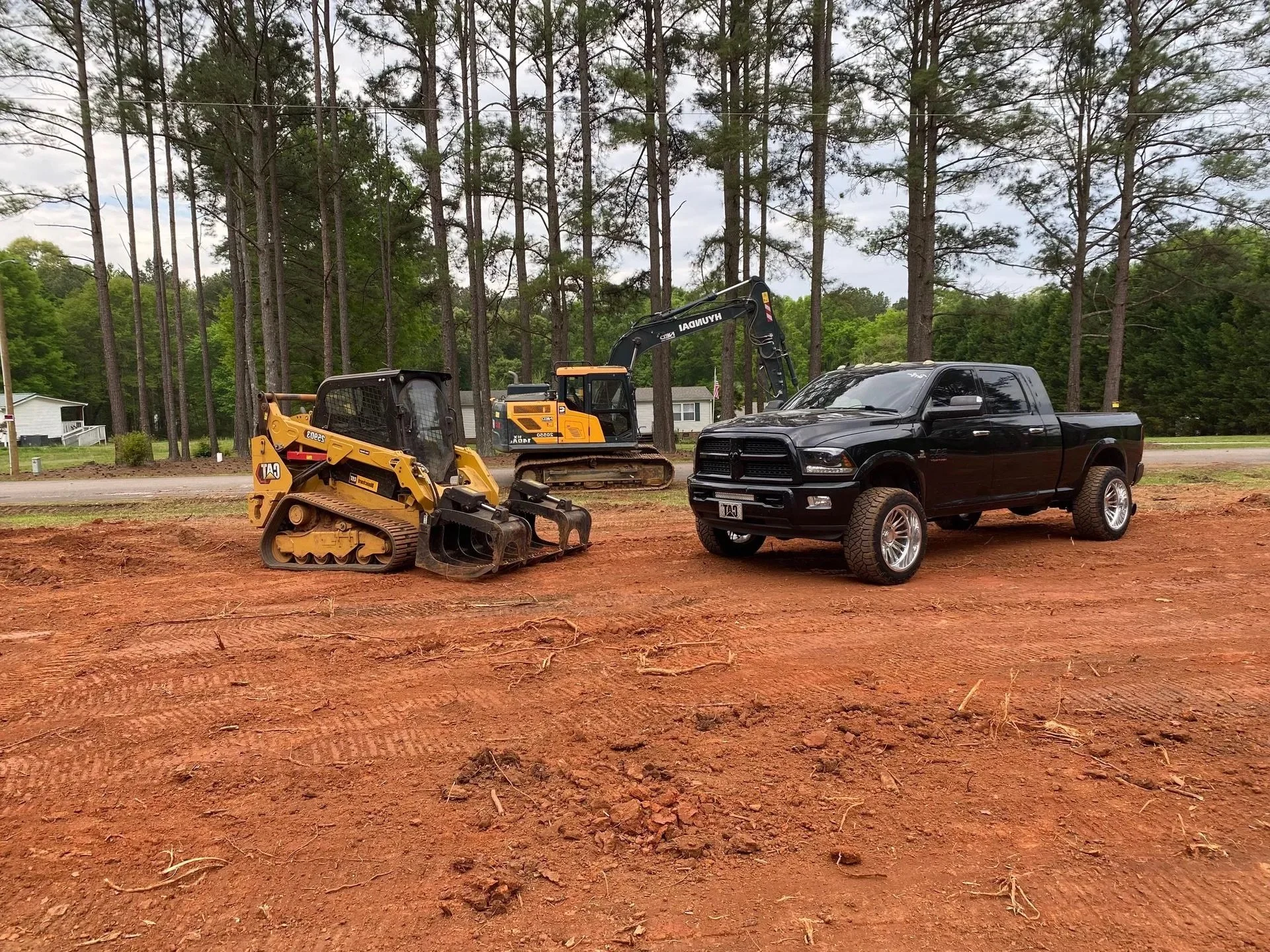 Construction equipment on a dirt lot with trees in the background, including a small bulldozer, a backhoe, and a black pickup truck.