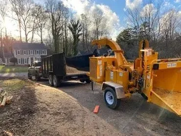 A large wood chipper attached to a truck, processing tree branches on a residential street in a neighborhood with trees and houses in the background.