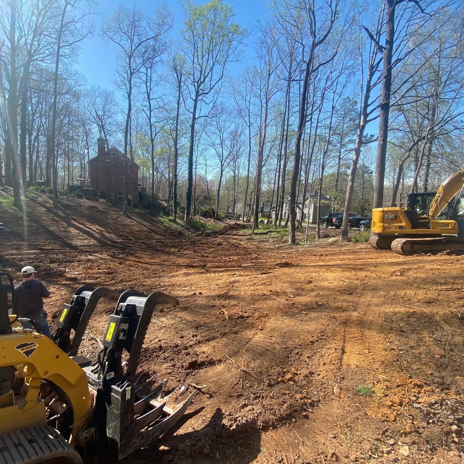 A construction site with a cleared dirt area, a yellow excavator on the right, and a person wearing a white helmet and dark shirt operating a small piece of machinery on the left. There are trees, some with green leaves, and houses in the background under a clear blue sky.
