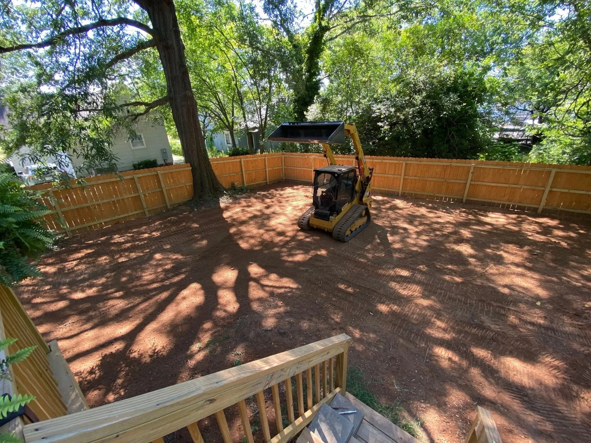 A backyard with a new wooden fence, a large tree, and a small excavator preparing the ground for landscaping.