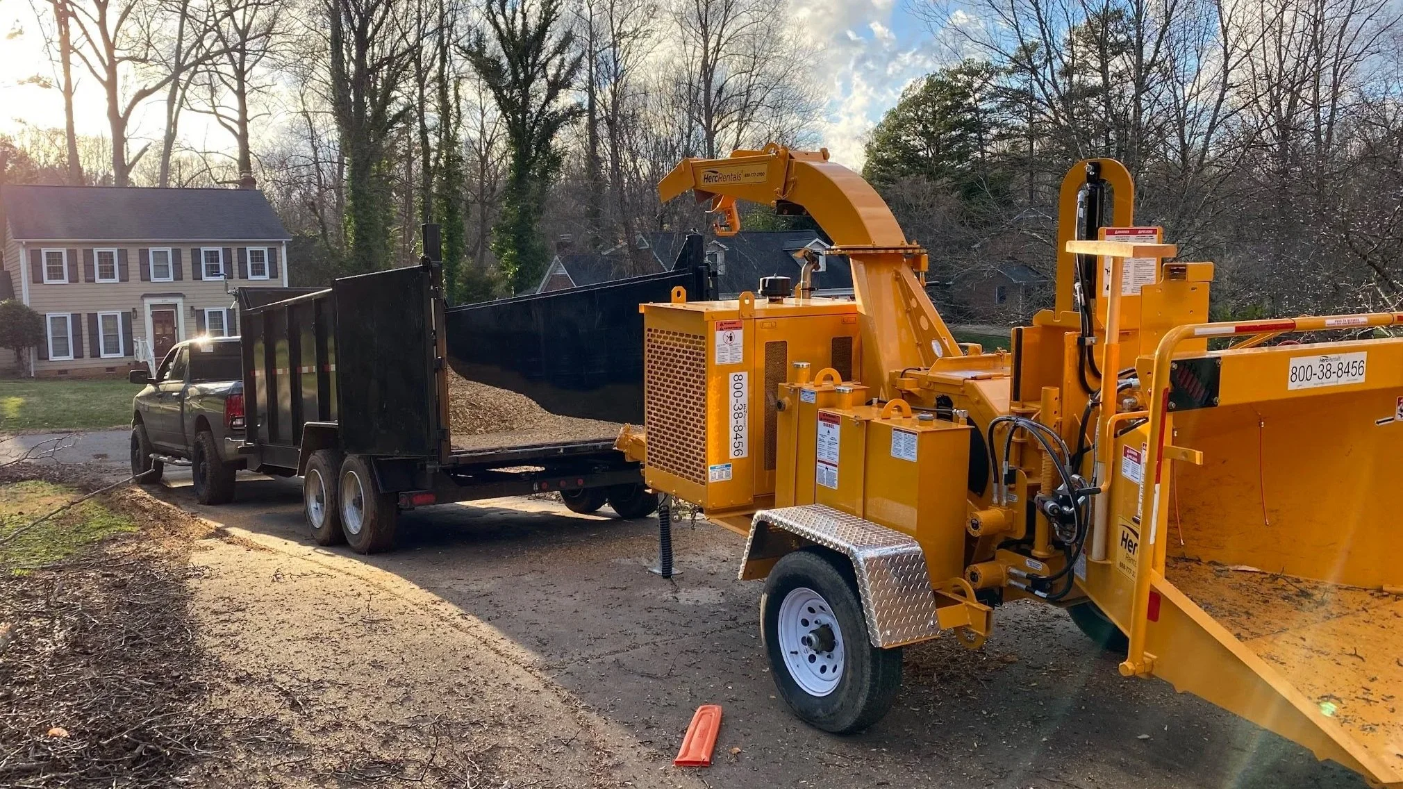 Construction equipment and a truck in a residential neighborhood during daylight, with houses and leafless trees in the background, and a dirt pathway in the foreground.