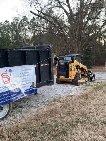 A small yellow excavator parked next to a black trailer on a grassy area near a gravel path and trees.