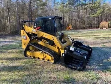 A yellow CAT compact track loader on grassy terrain with trees in the background.