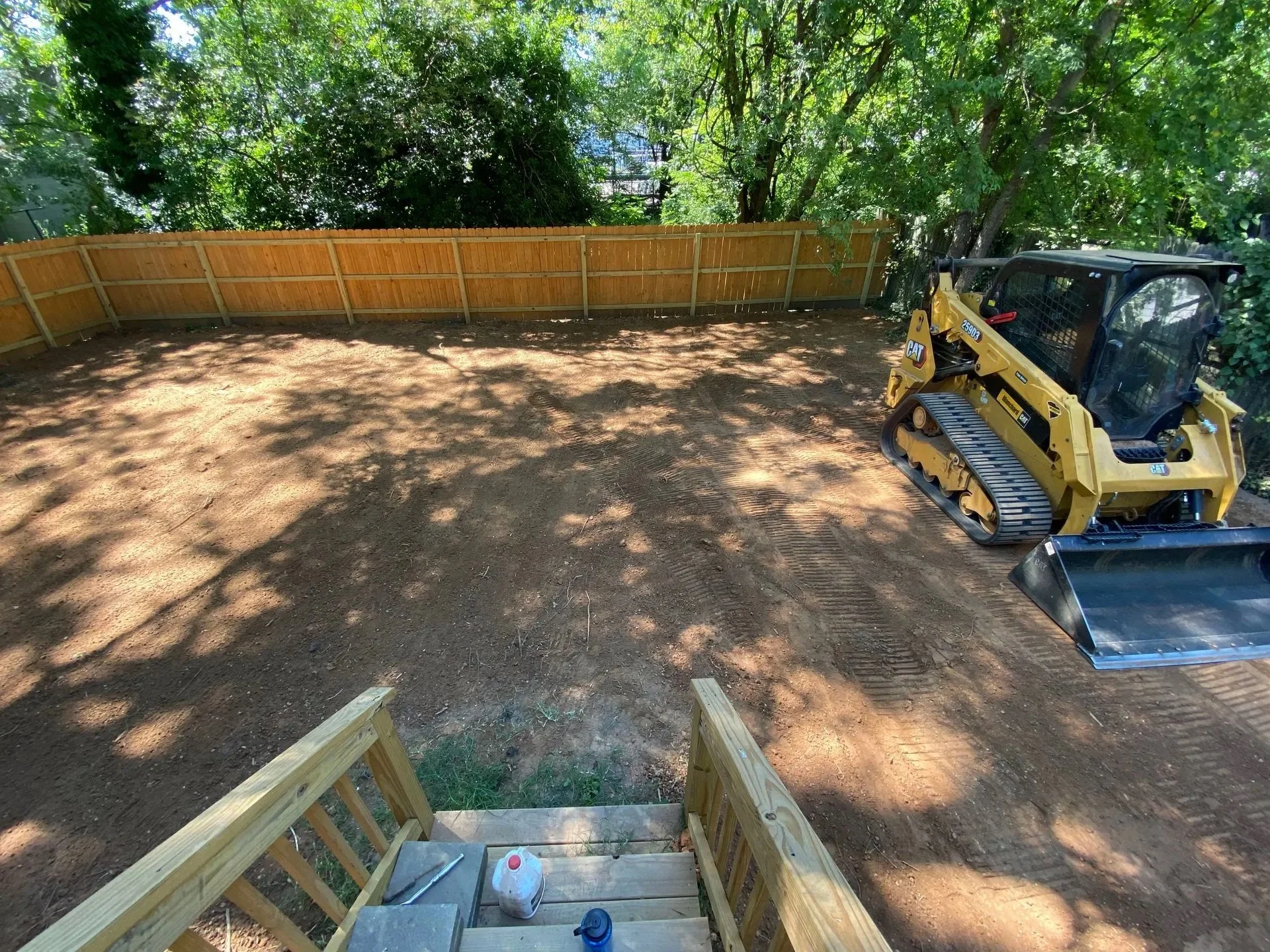 A backyard under construction with a dirt ground, a yellow CAT mini excavator, and a wooden fence along the perimeter. A set of wooden stairs is leading down into the yard.