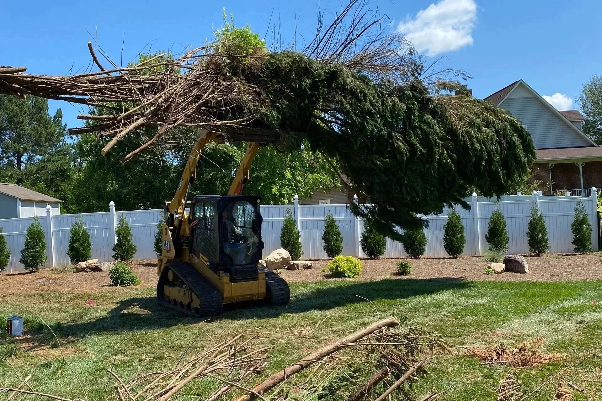 A small construction vehicle lifting a large tree branch over a backyard with a white fence and small bushes.