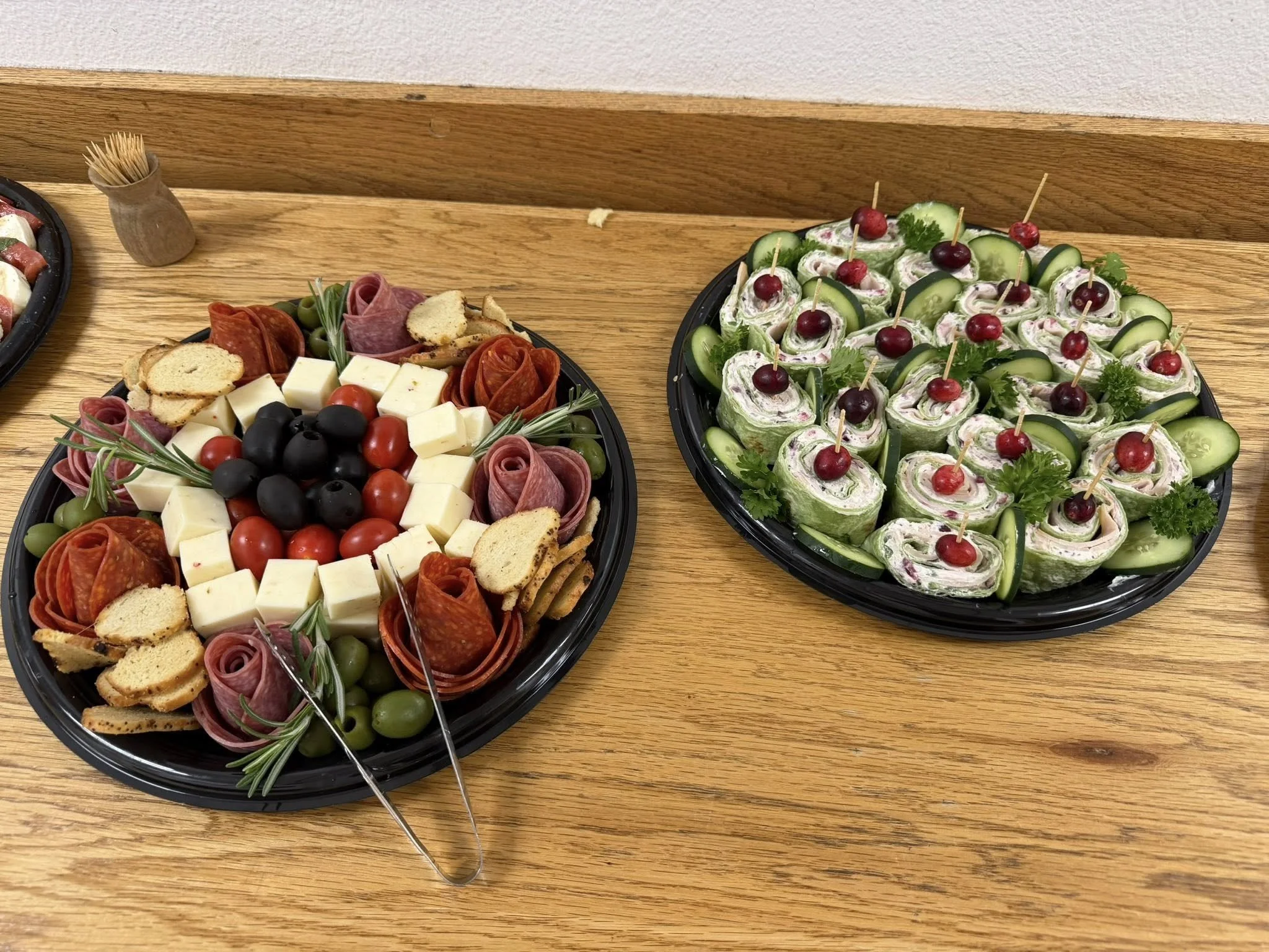 Platters of cheese, olives, meats, cherry tomatoes, and vegetable roll-ups on a wooden table.