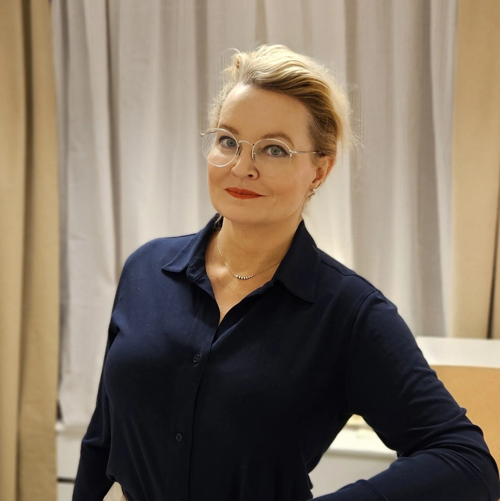 En kvinna med glasögon och blont hår, klädd i en mörkblå skjorta, står framför ett gardinlöst fönster.