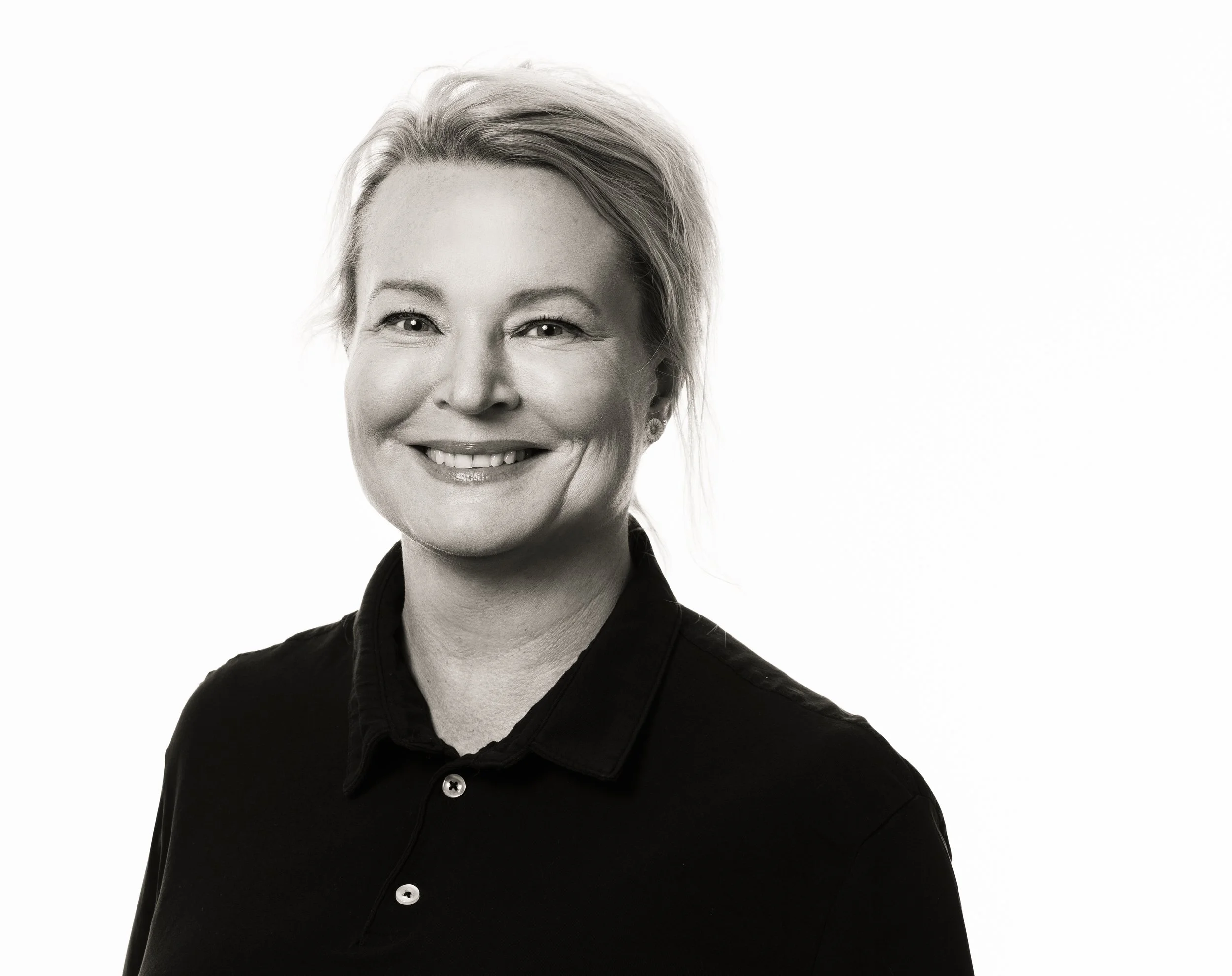 Svartvitt porträtt av en kvinna med blond hår, leende och bärande en mörk skjorta, mot en vit bakgrund.