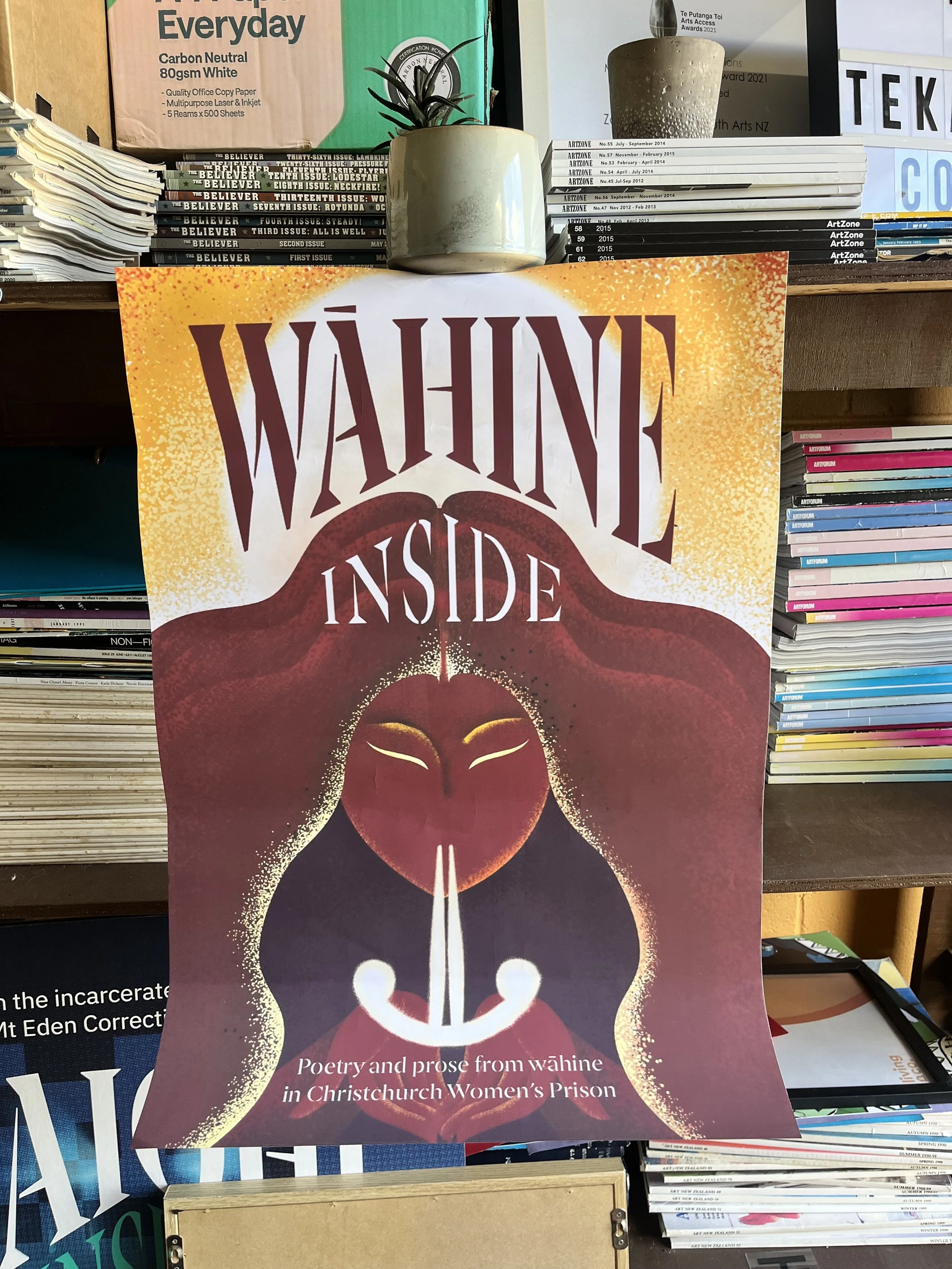 A poster titled "Wāhine Inside" with artwork of a woman with long red hair and closed eyes, holding a smiley face with a line of white smoke coming from its top, representing poetry and prose from wāhine in Christchurch Women's Prison. The poster is displayed among stacks of magazines and papers.