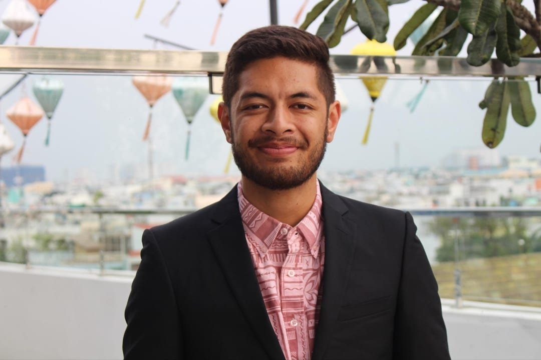 A young man with short dark hair and a beard, wearing a black blazer and pink patterned shirt, standing outdoors on a rooftop with a cityscape in the background and decorative hanging glass objects above.