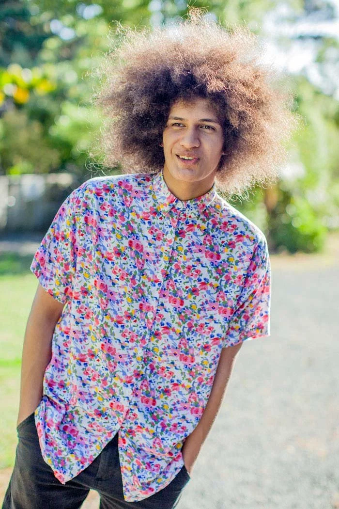 A young man with curly  hair wearing a colourful, floral short-sleeve shirt, standing outdoors in a sunny environment with green trees in the background.