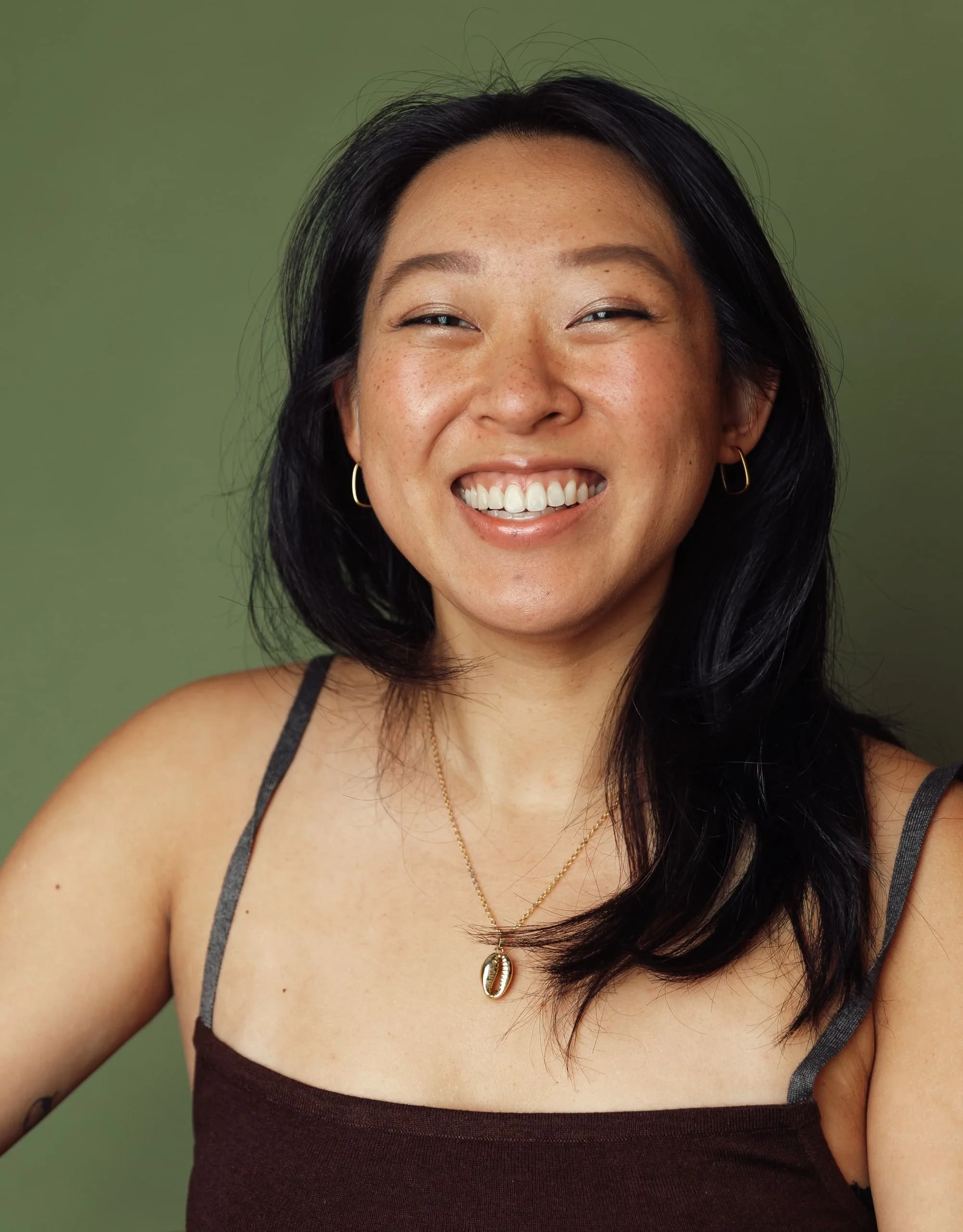 A smiling woman with black hair, wearing gold hoop earrings and a necklace with a shell pendant, posing against a green background.