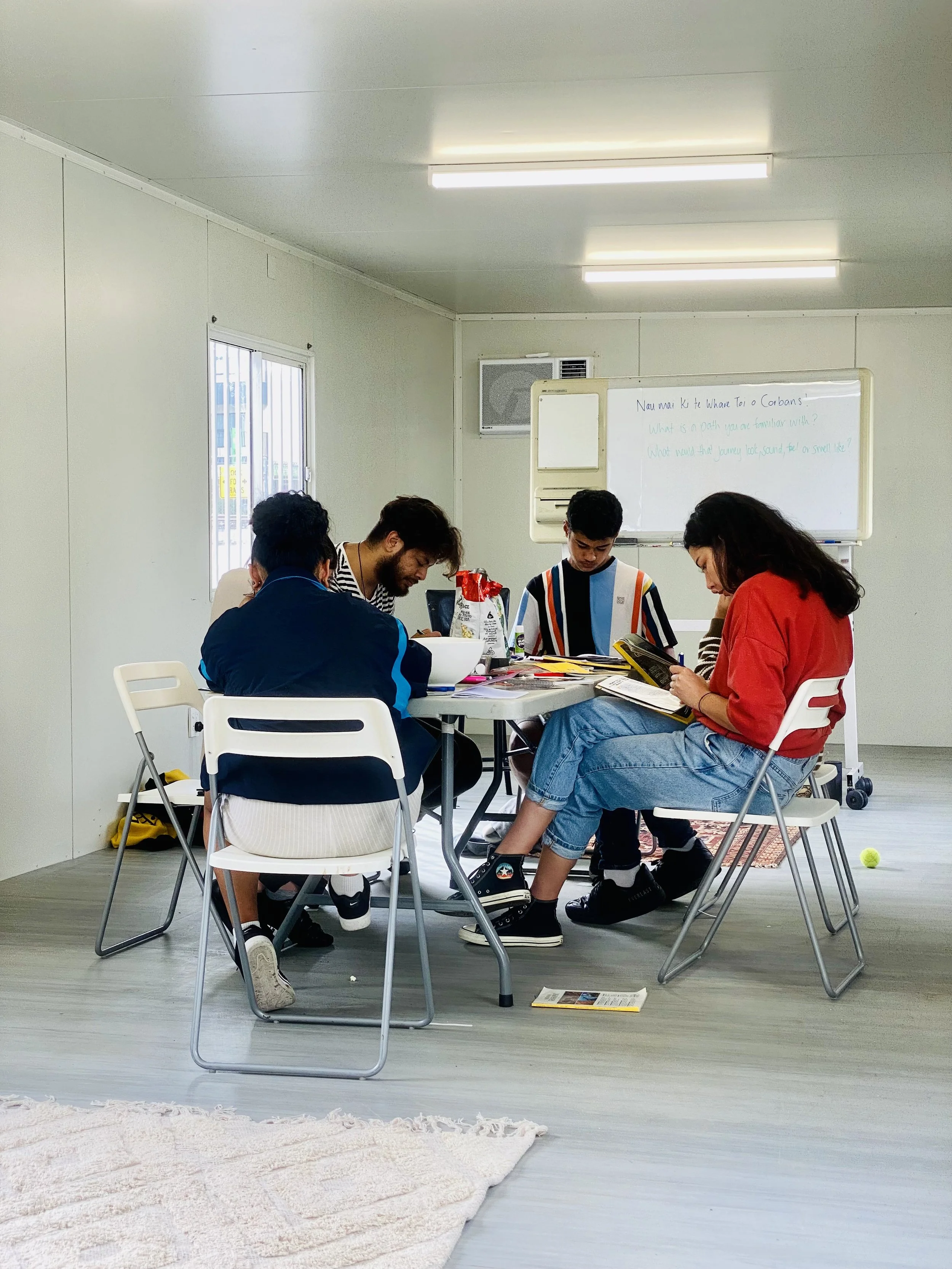 Group of five young adults sitting around a table in a plain room, engaged in studying or discussion, with books and snacks on the table. Whiteboard in the background with writing, and a window on the left.