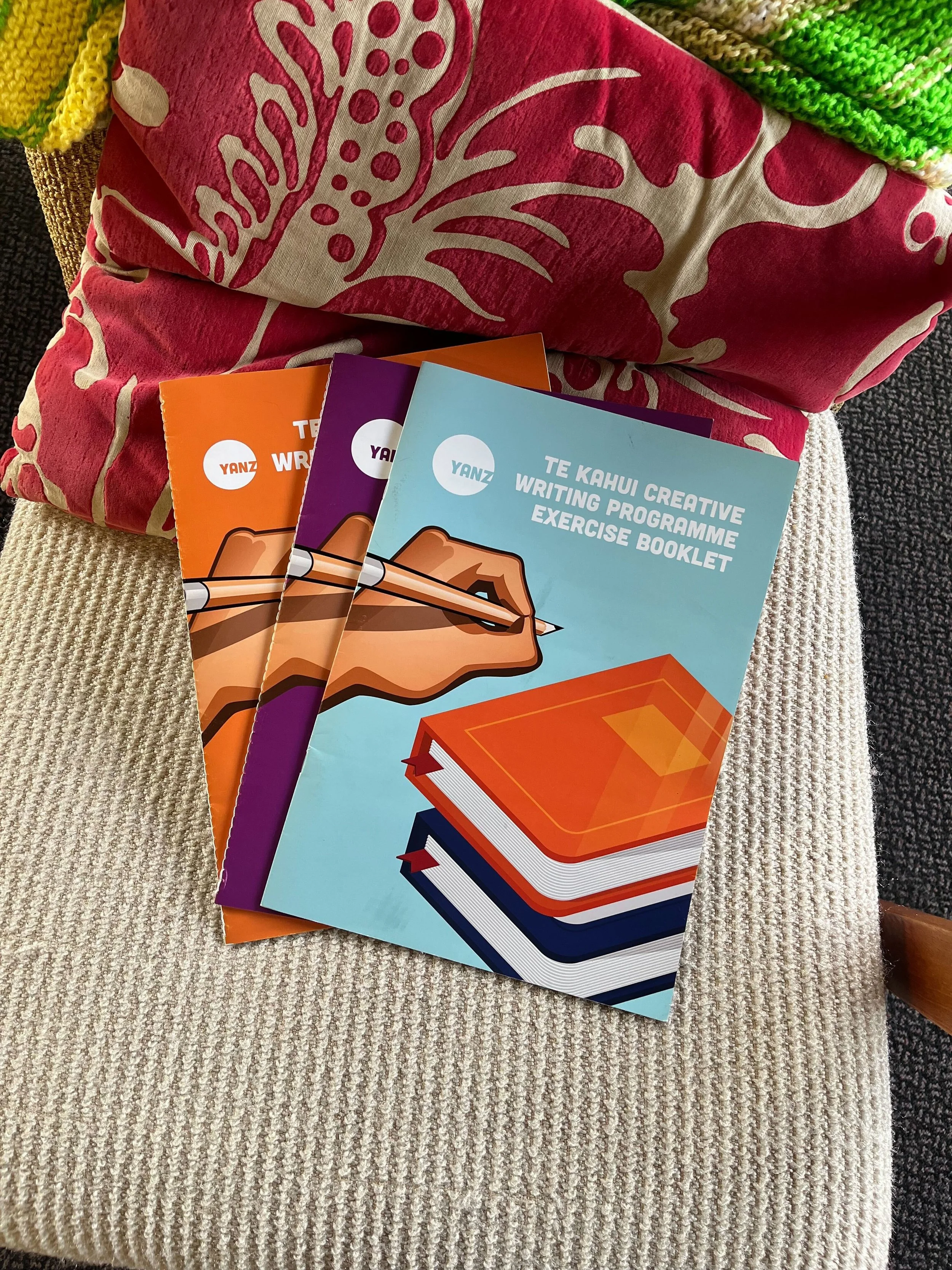 A stack of three colorful booklets on a beige, textured fabric surface. The booklets are titled 'Te Kahul Creative Writing Programme Exercise Booklet.' The cover of the front booklet features an illustration of a hand holding a pencil, writing on a stack of books, with a light blue background. There are pink, orange, purple, and blue covers visible.