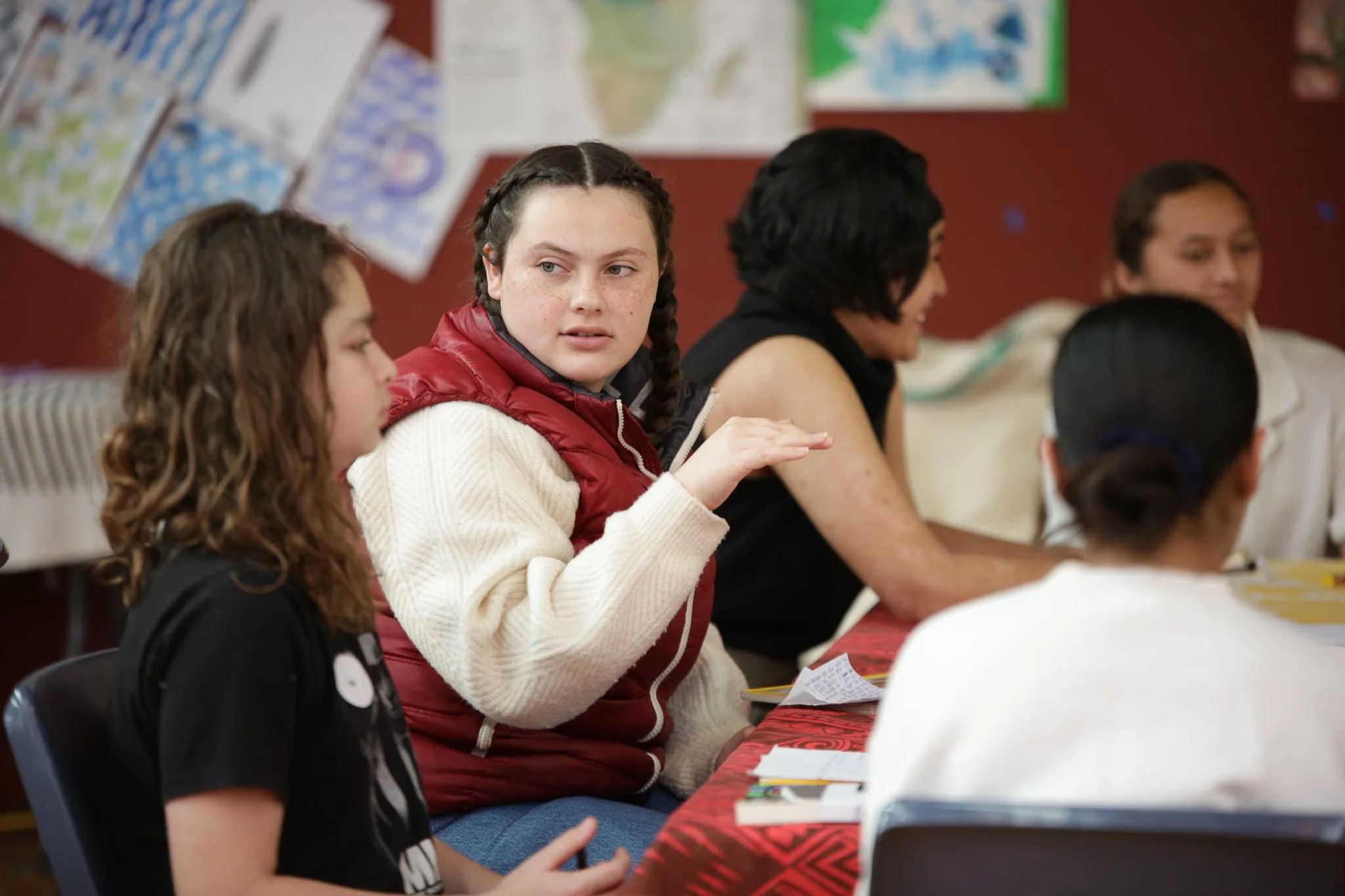 A group of diverse young people sitting at a long table engaged in conversation or an activity, with colorful posters or artwork on the wall behind them.