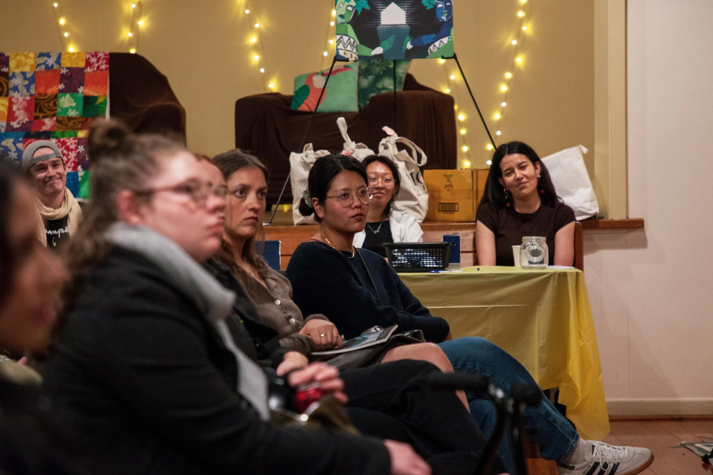 Group of people seated and listening at an indoor event, with fairy lights and gifts in the background.