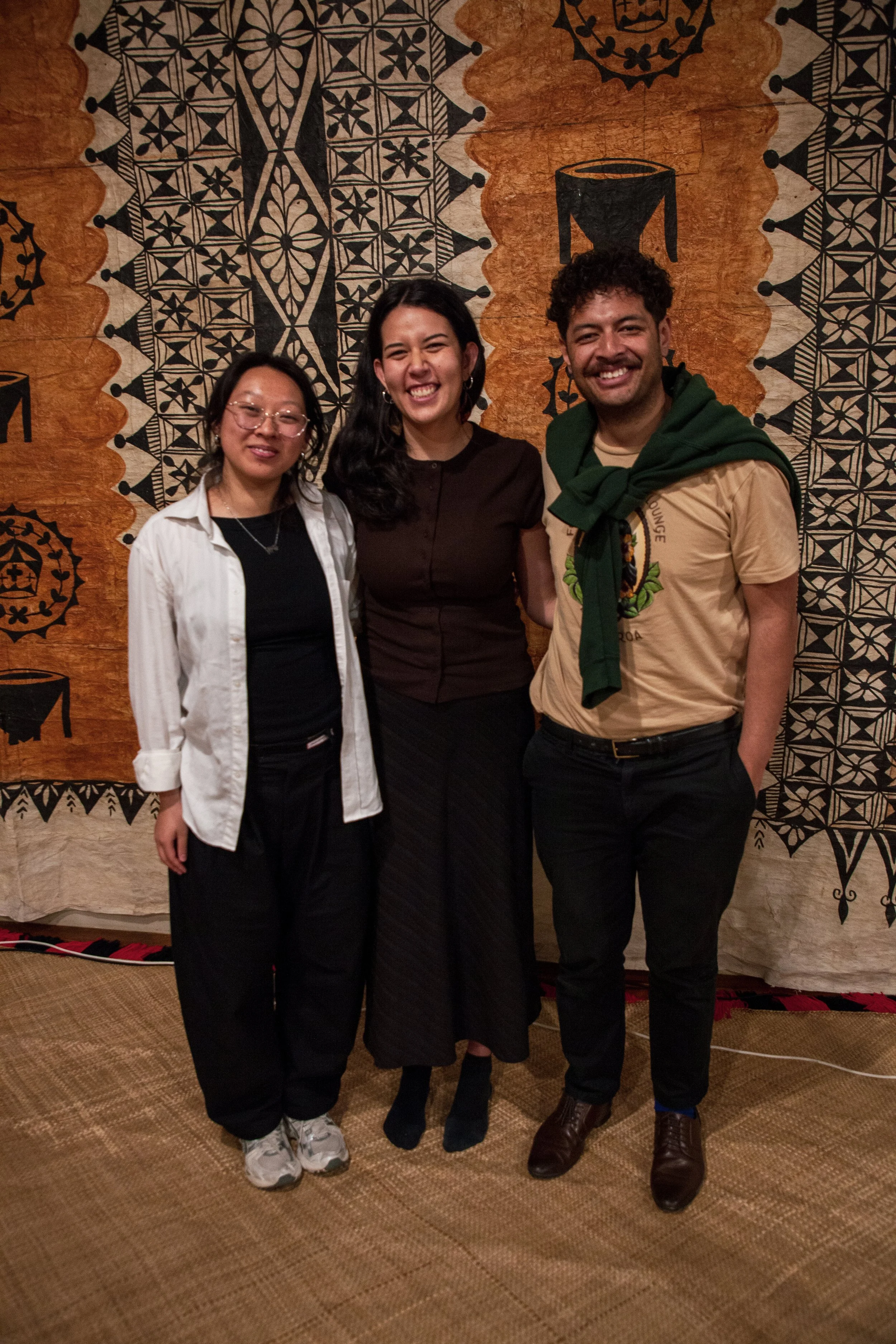 Three people standing together indoors in front of a patterned wall, smiling at the camera.