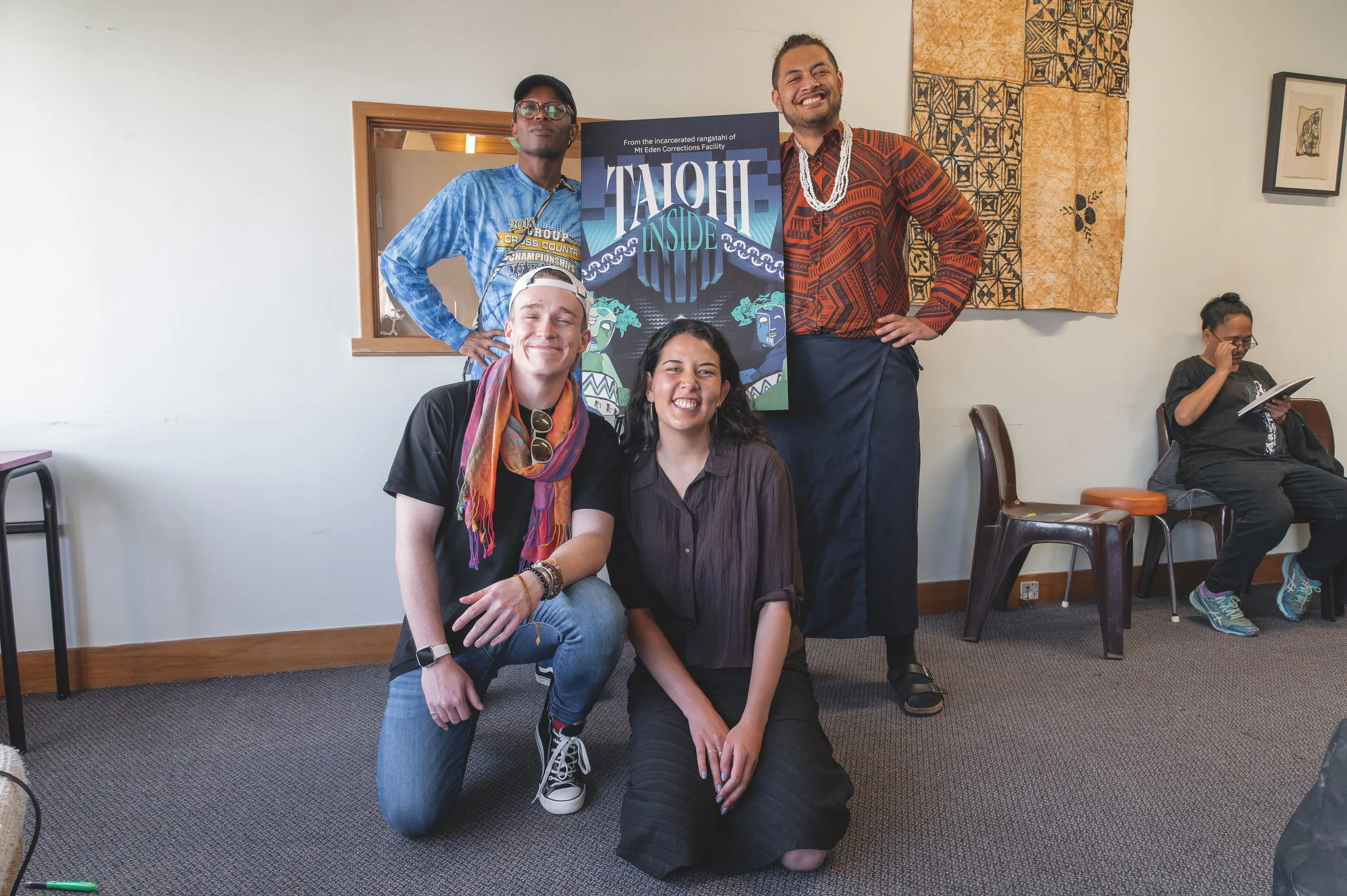 Five people posing in a room, three standing in the back and two kneeling in front, smiling for the photo. One woman is sitting on the floor, and a woman is sitting on a chair, reading a book. There is a sign in the background that reads "TAIOHI INSIDE" and some artwork on the wall.