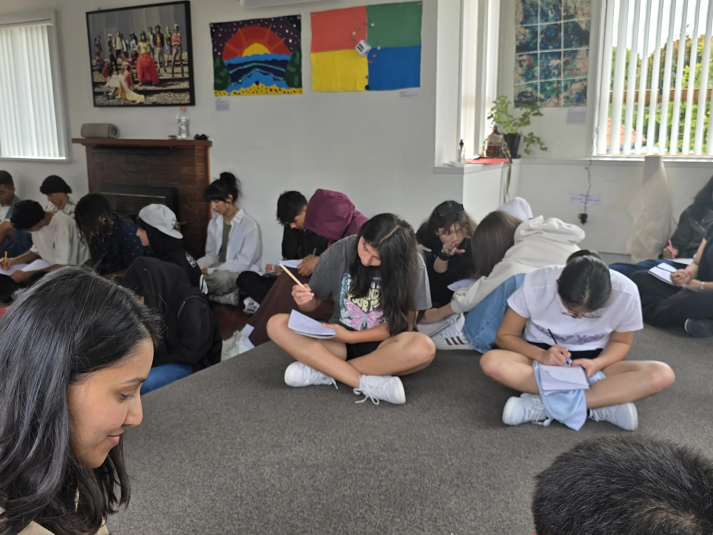 A group of young students sitting cross-legged on the floor in a classroom, engaged in note-taking or writing activities, with colorful paintings and artworks displayed on the wall behind them.