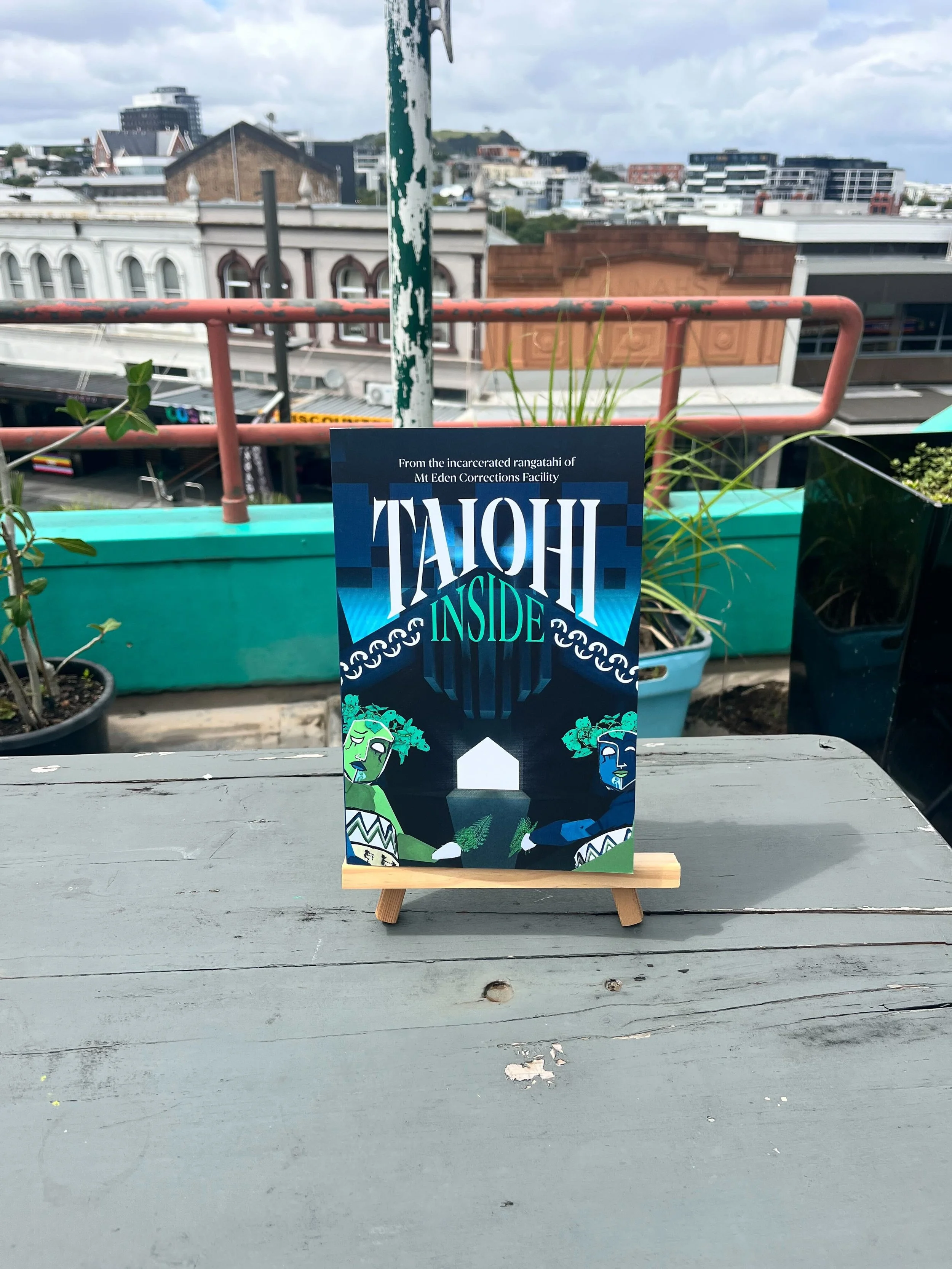 A colorful brochure or sign on a small easel on a gray wooden table, with a cityscape background, featuring the title 'TAYOH INSIDE' and illustrations of two stylized faces, set against a dark abstract background.