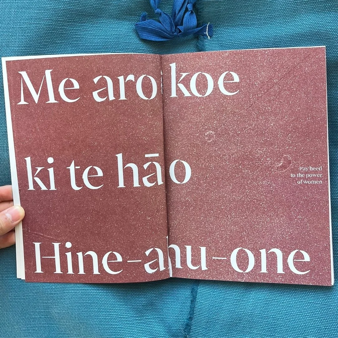 Open book with large white text on pink background, phrases in Maori language, and small white text in English reading 'Pay heed to the power of women,' on a teal fabric surface.