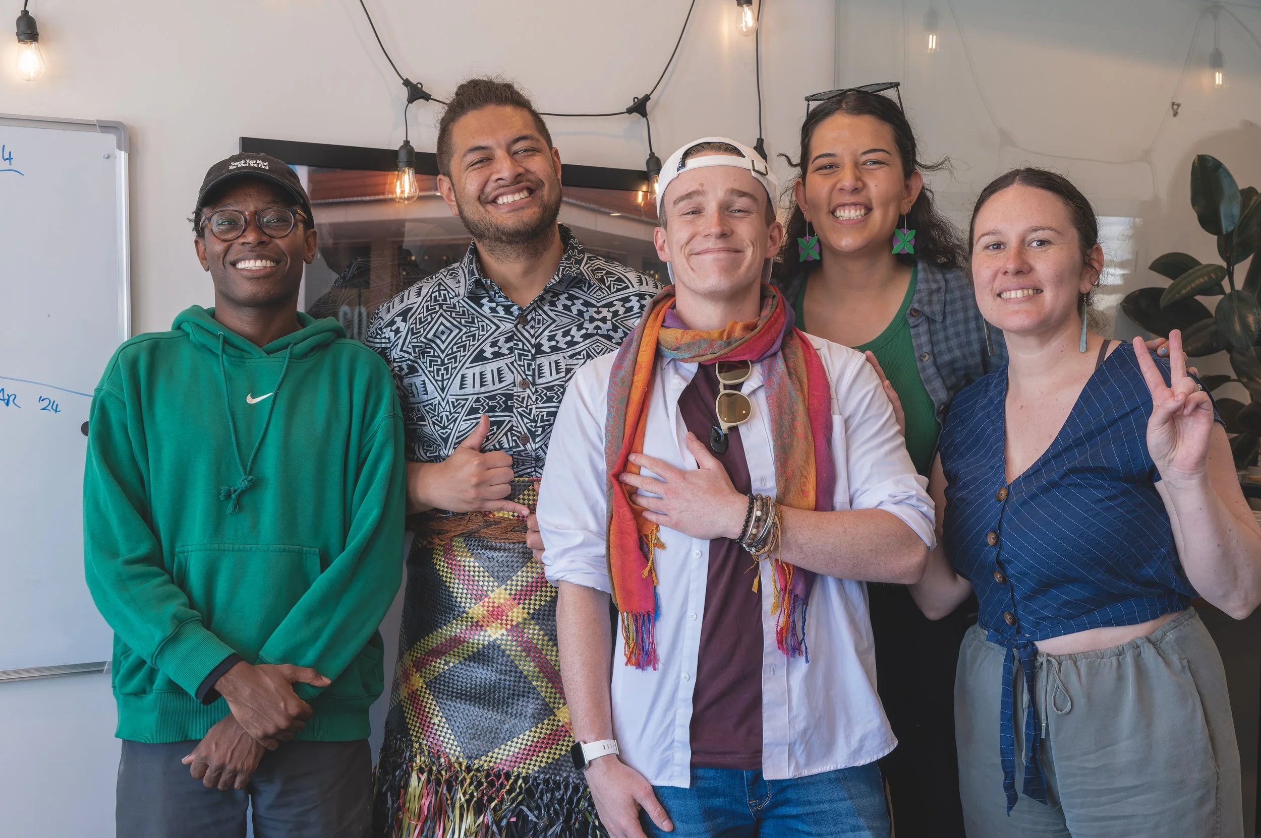 Group of five diverse young adults smiling and posing for a photo indoors, with string lights hanging from the ceiling and a whiteboard in the background.
