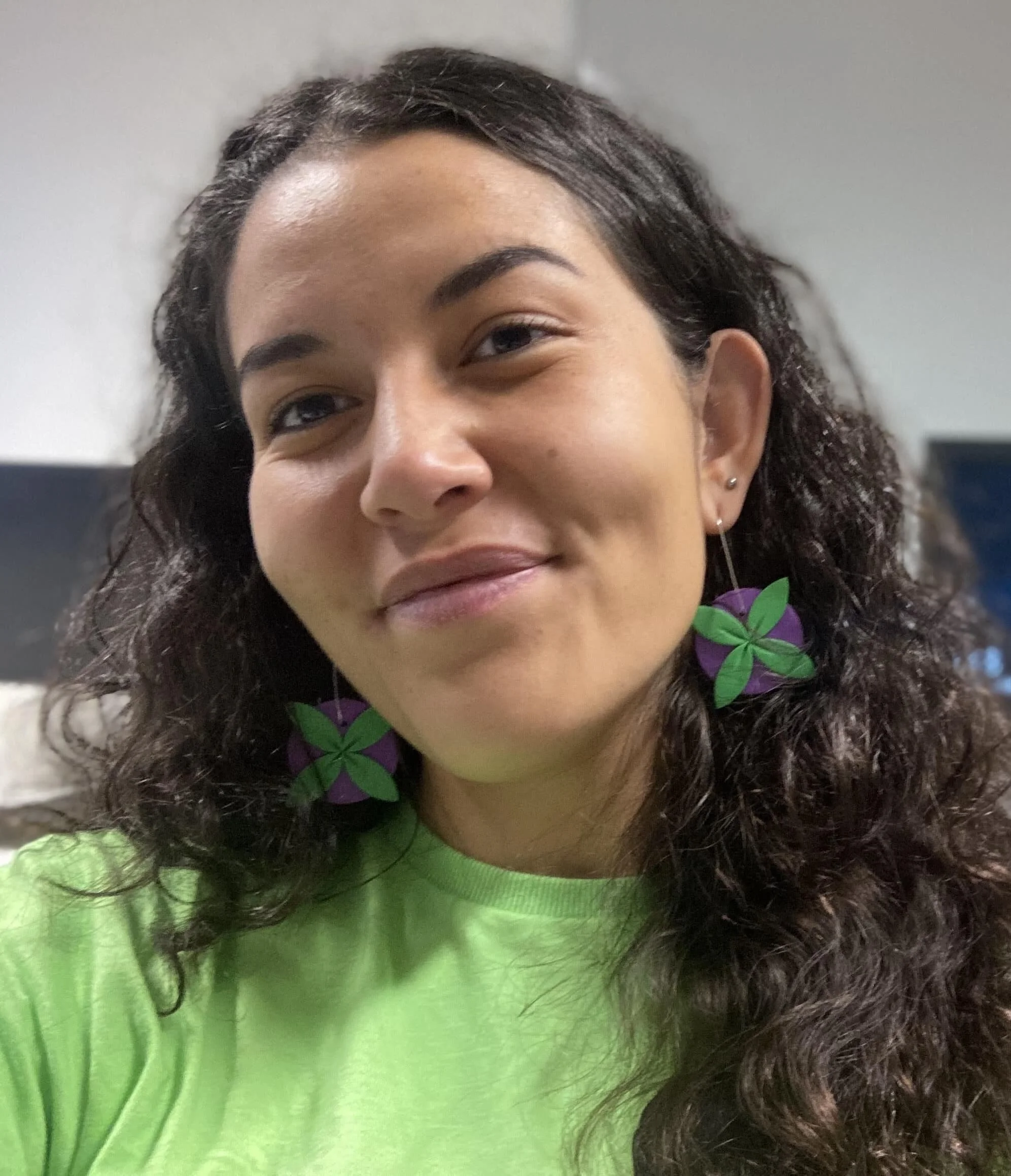 A woman with long curly dark hair wearing large purple and green flower-shaped earrings and a bright green shirt, smiling softly.