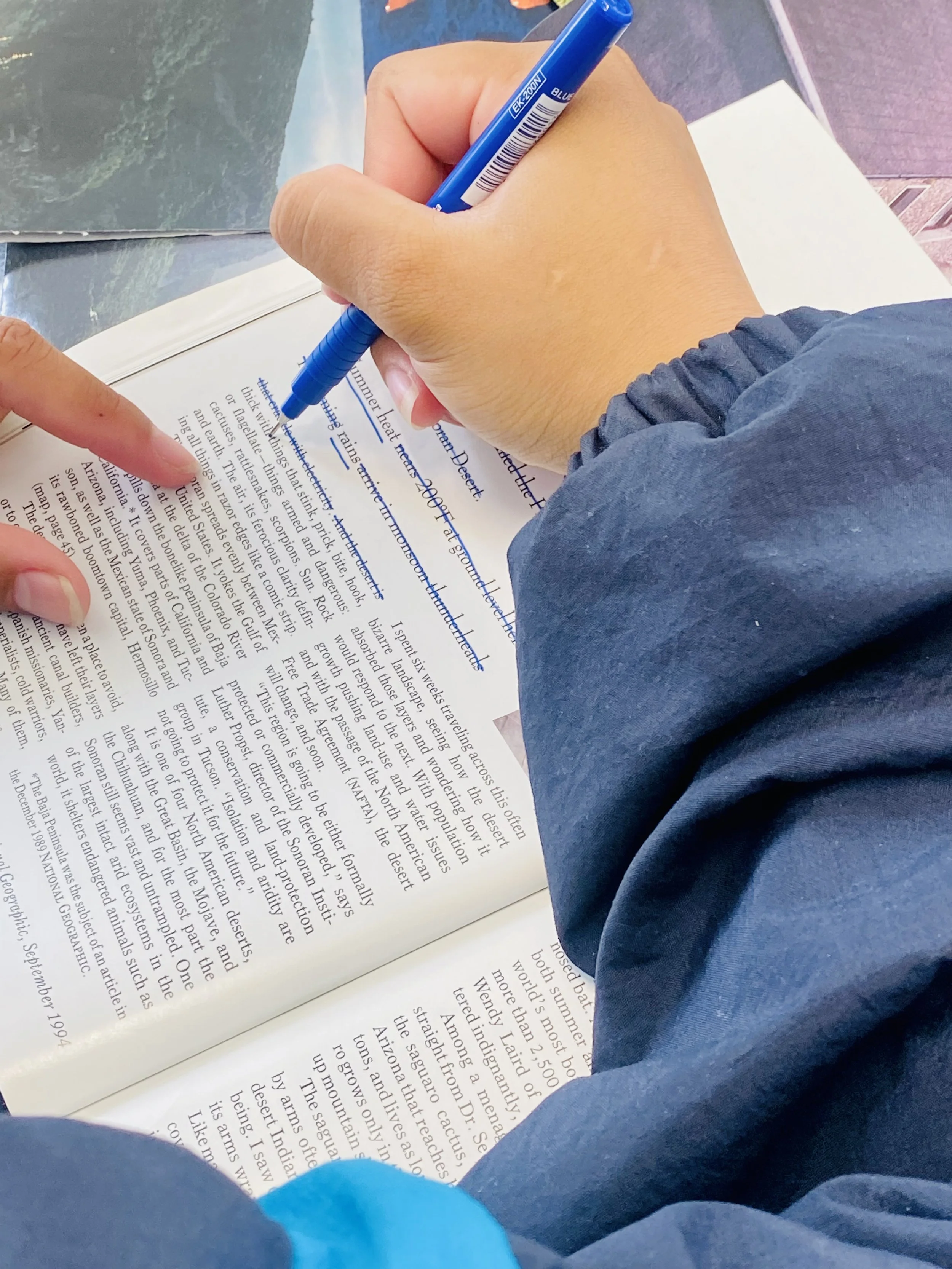 Person reading a book and pointing at a sentence with their finger while taking notes with a blue pen.