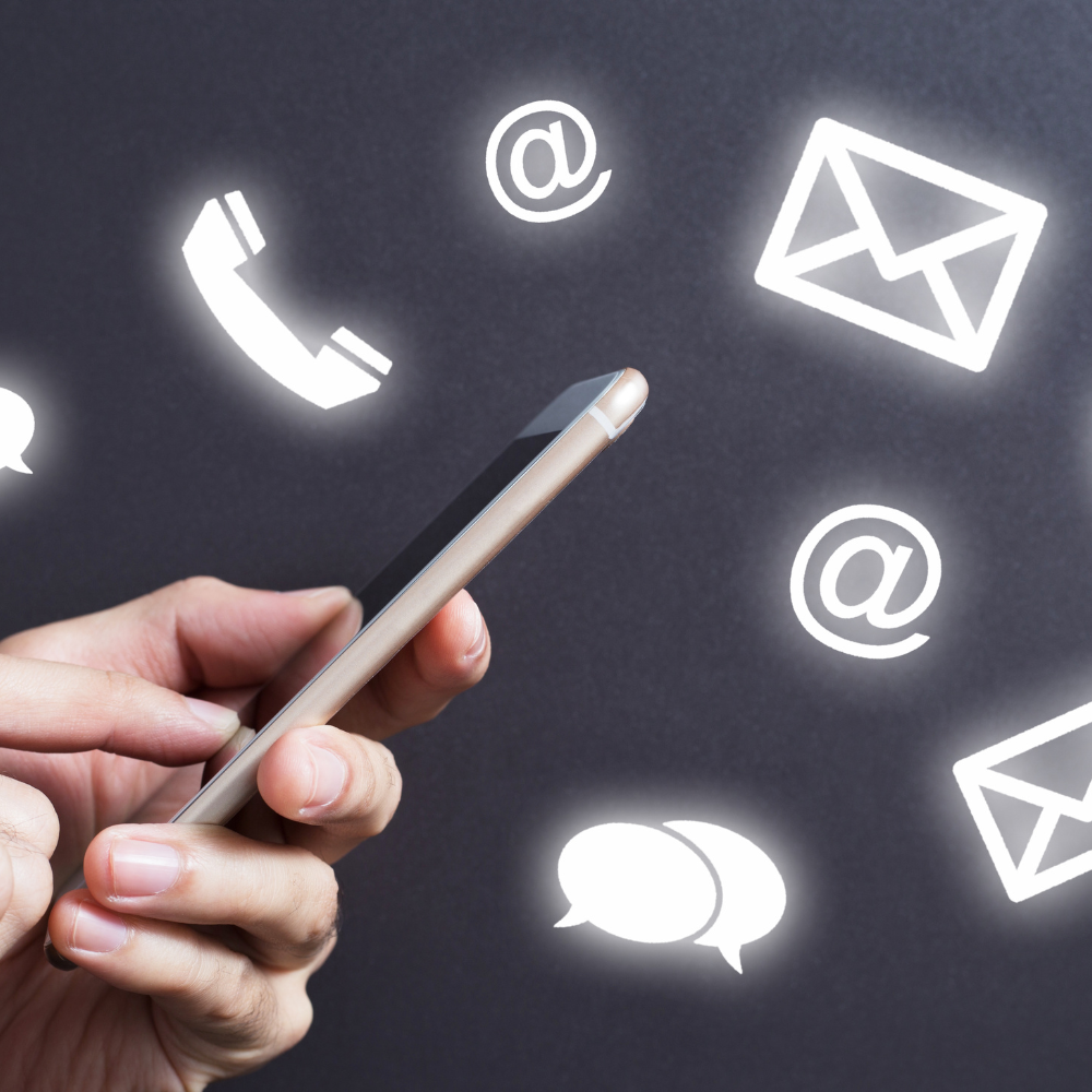 Person holding a smartphone with glowing icons of a phone receiver, email envelopes, the at symbol (@), and speech bubbles floating around against a dark background.